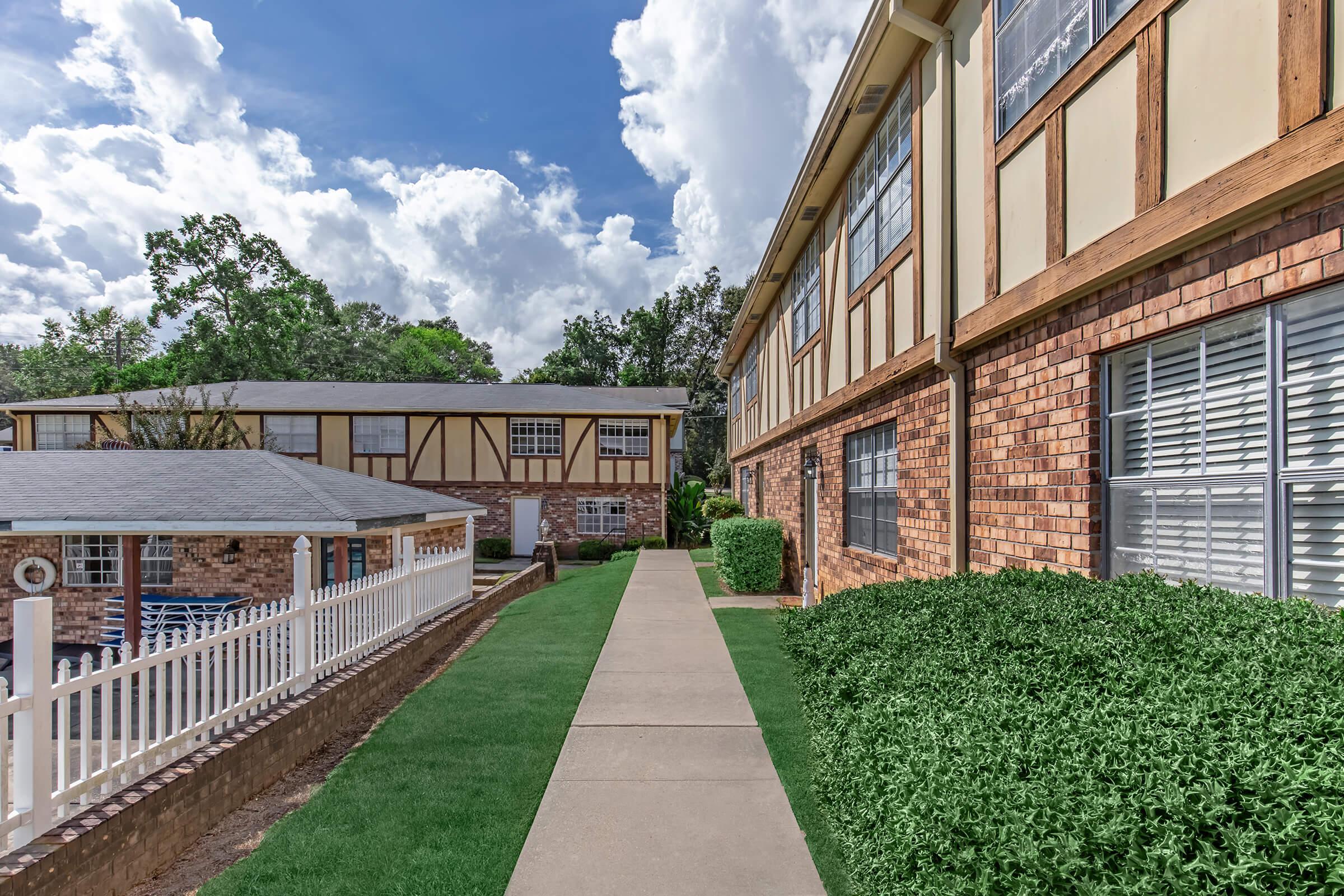 A pathway leading between two brick buildings with a mix of traditional and modern architecture. Lush green grass borders the walkway, and a swimming pool is visible to the left, with white fencing. The sky is partly cloudy, adding to the bright, inviting atmosphere.