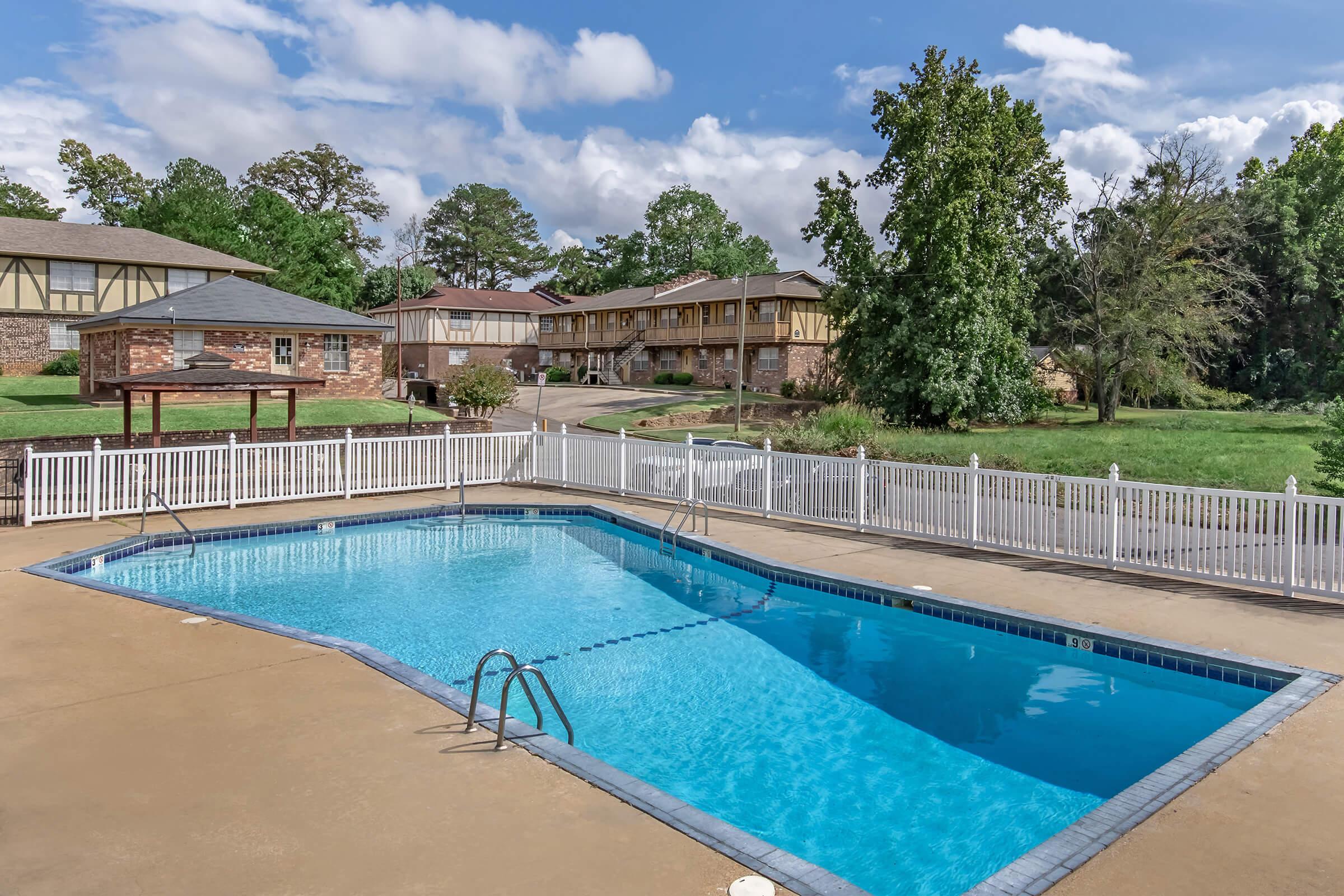 A clear blue swimming pool surrounded by a white fence, with a gazebo nearby. In the background, there are several brick and wooden buildings under a bright sky with scattered clouds. Lush greenery surrounds the area, creating a relaxing atmosphere.