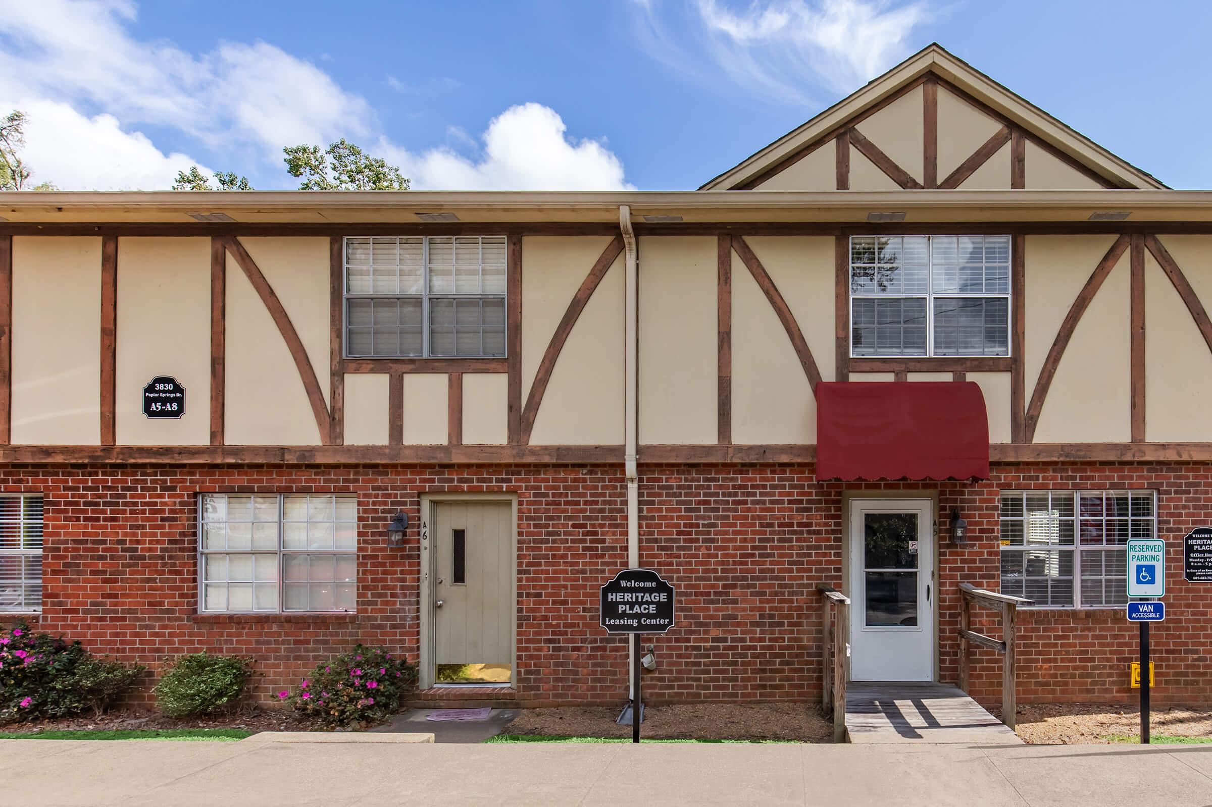 Two-story residential building featuring a combination of brick and siding. The front has a red awning over the entrance, with a ramp for accessibility. Flower beds on either side of the door and a sign indicating the "Heritage Place Leasing Center" are visible. Blue sky with scattered clouds above.