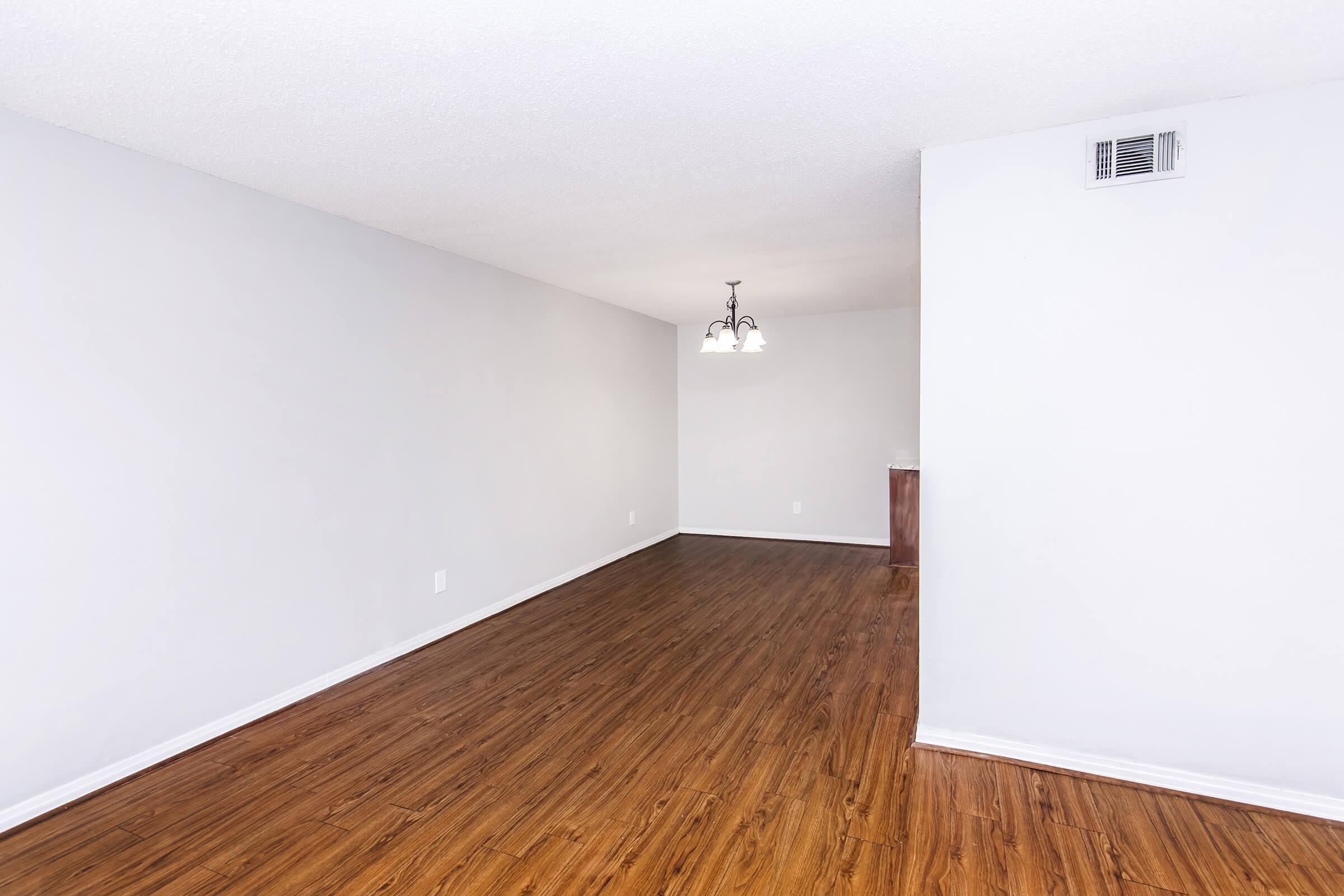 A spacious, empty room with light-colored walls and dark wooden flooring. A simple light fixture hangs from the ceiling, and a small kitchen area is visible in the background. The space has a minimalist aesthetic, making it suitable for various furnishings and decor styles.