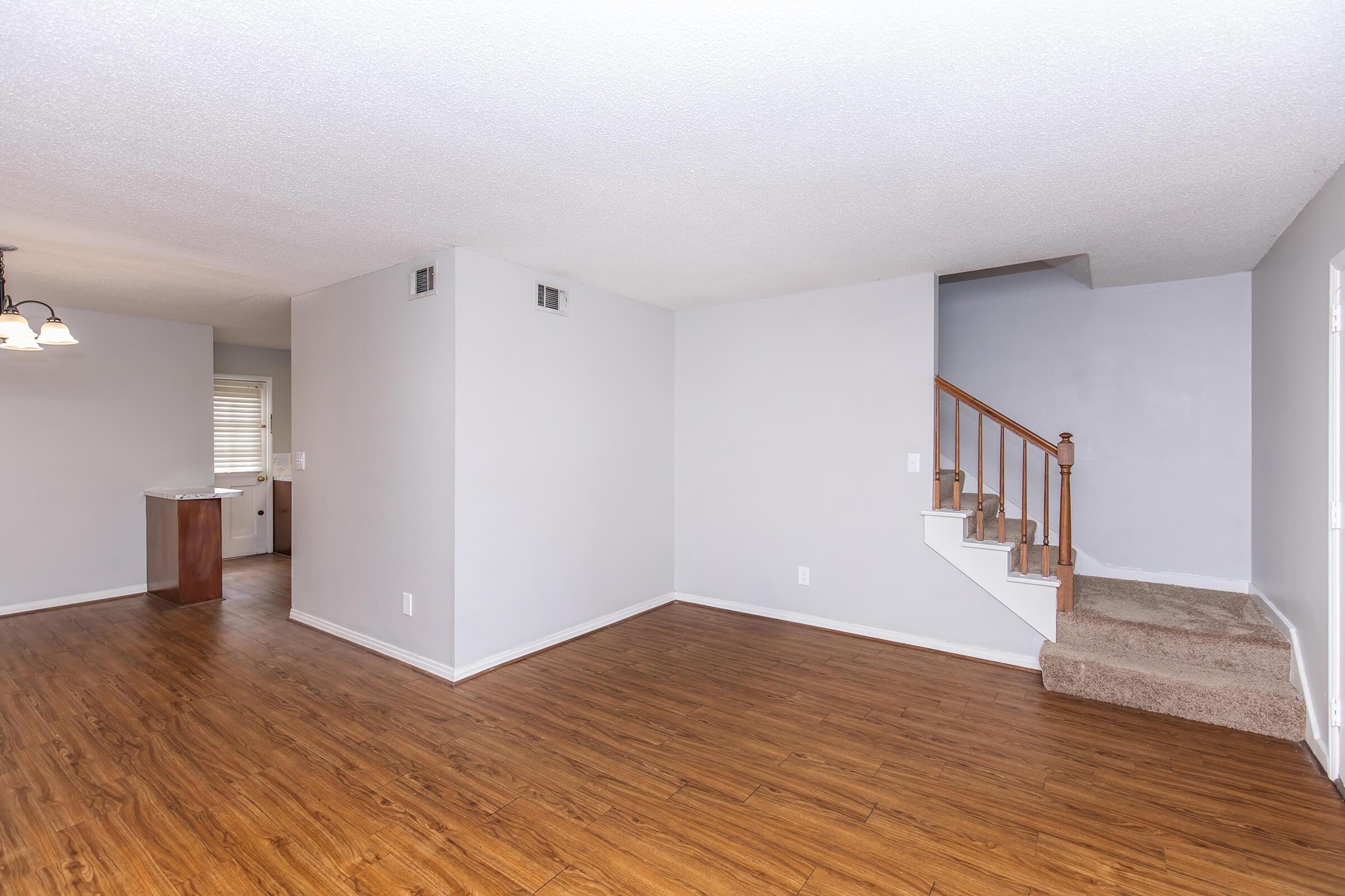Interior of a modern living space featuring light gray walls and hardwood-style flooring. A staircase is visible on the right leading to an upper level. To the left, there's a glimpse of a kitchen area with a small dining space. Natural light enters through a window, creating a bright and open atmosphere.