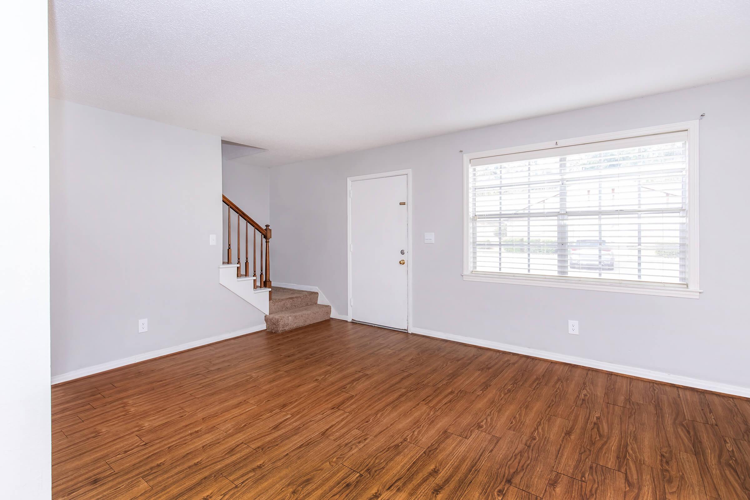 Empty living room featuring light gray walls, a wooden staircase on the left, and a large window with white blinds. The floor is covered in brown laminate, and there is a plain white door in the back. The space has an open and airy feel with natural light coming in.