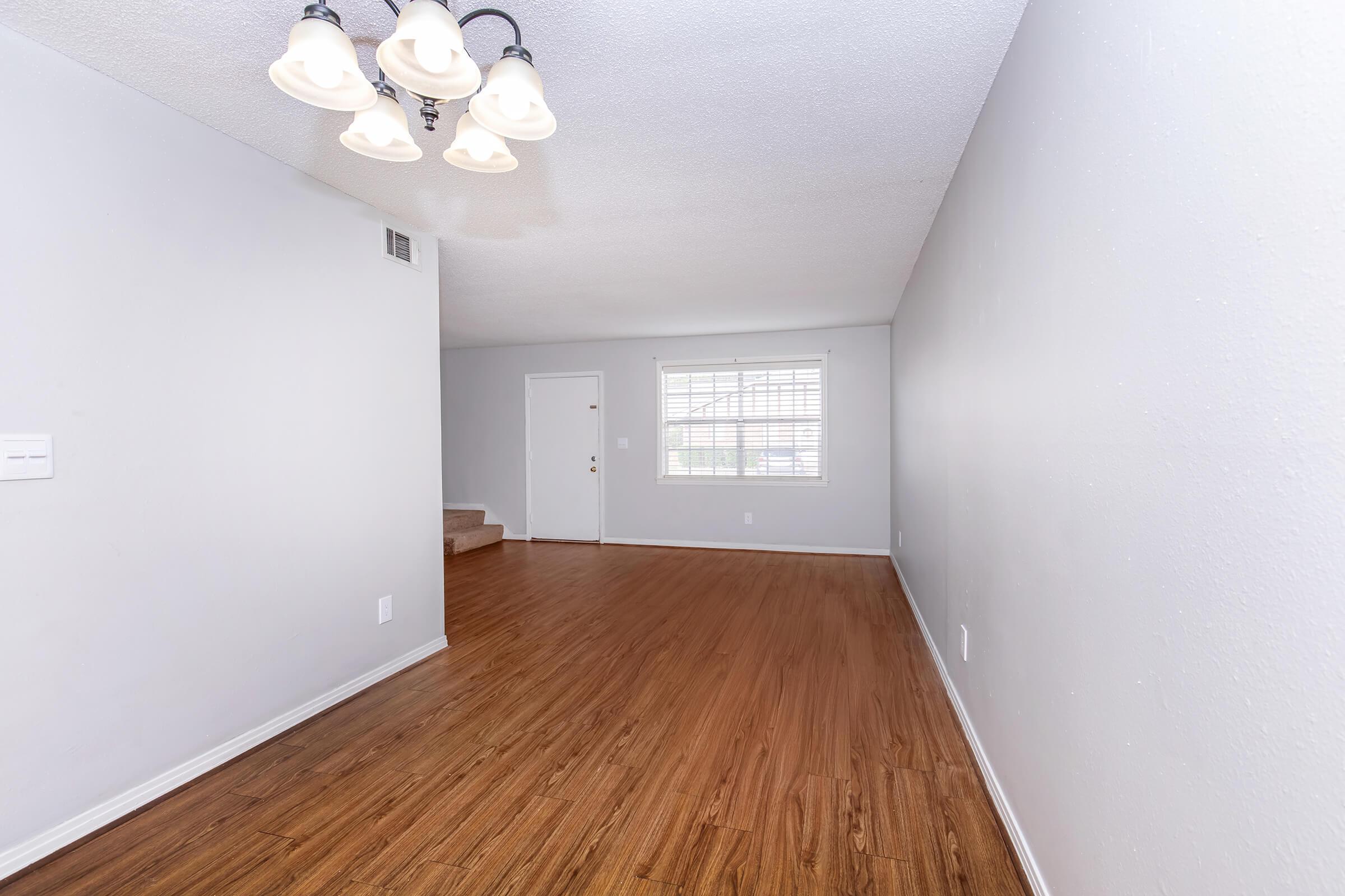 Spacious interior of a living area featuring light-colored walls, hardwood flooring, and a ceiling light fixture. The room includes a doorway leading outside, a window with blinds, and a staircase in the background. The space is bright and open, suitable for various furniture arrangements.