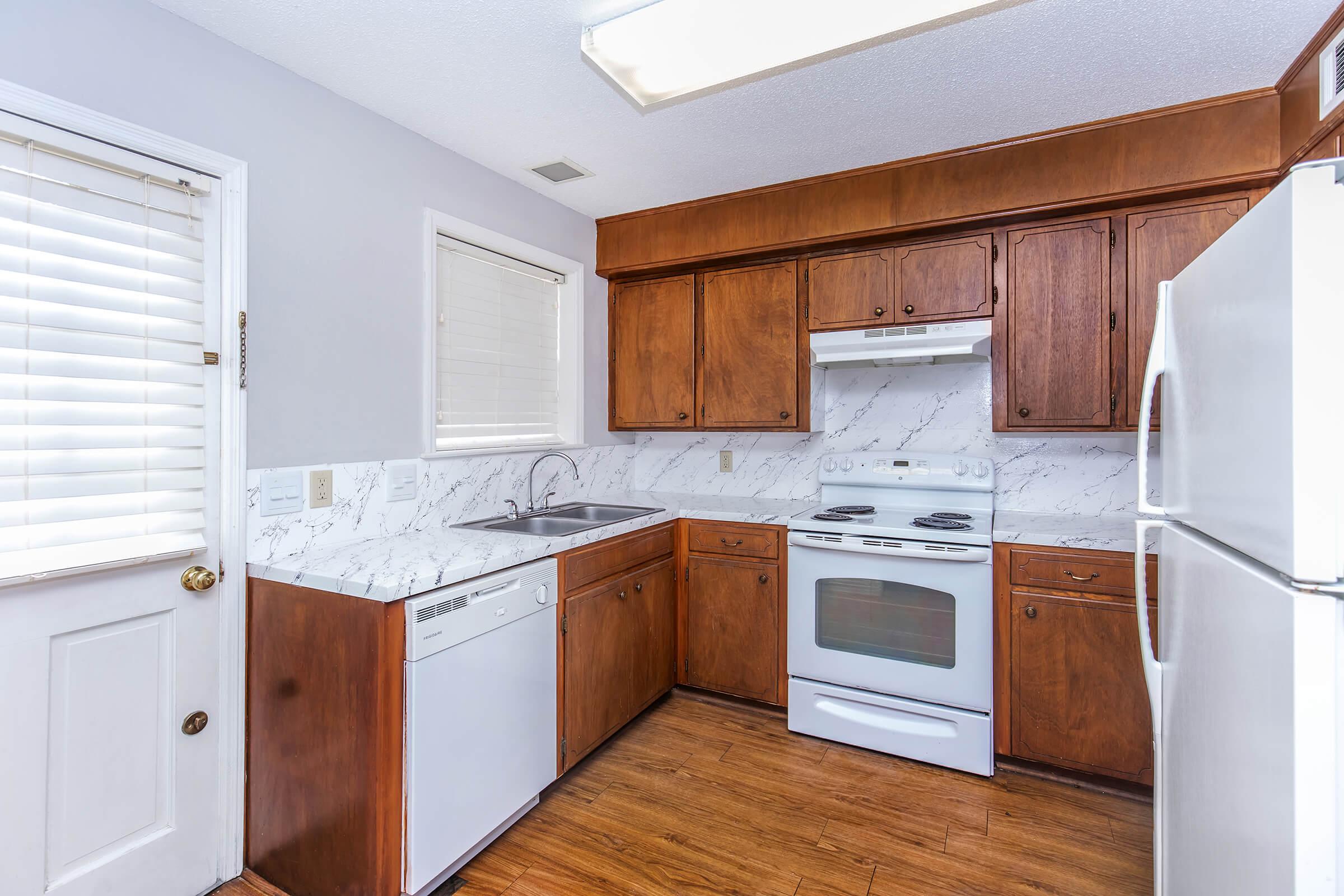 Kitchen featuring wooden cabinets, a white stove, and a sink with a dishwasher. The countertops have a marble-like finish, and there is a white refrigerator. Natural light enters through a window and a door with blinds. The floor is covered with wood-like tiles.