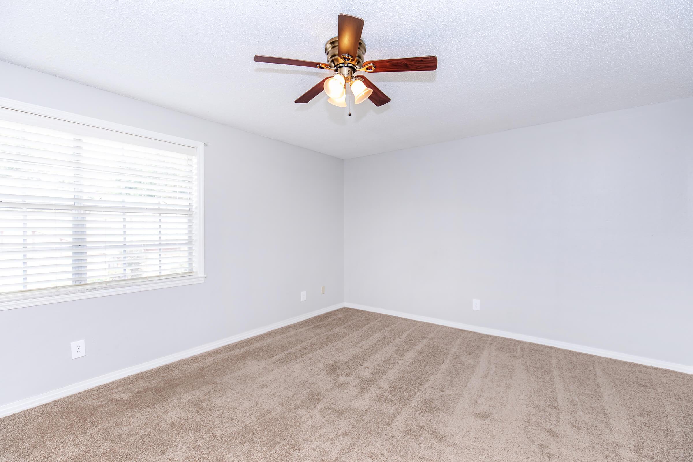A light-colored empty room featuring beige carpet, a ceiling fan with light fixtures, and a window with white blinds allowing natural light. The walls are painted in a light shade, creating a bright and open atmosphere.
