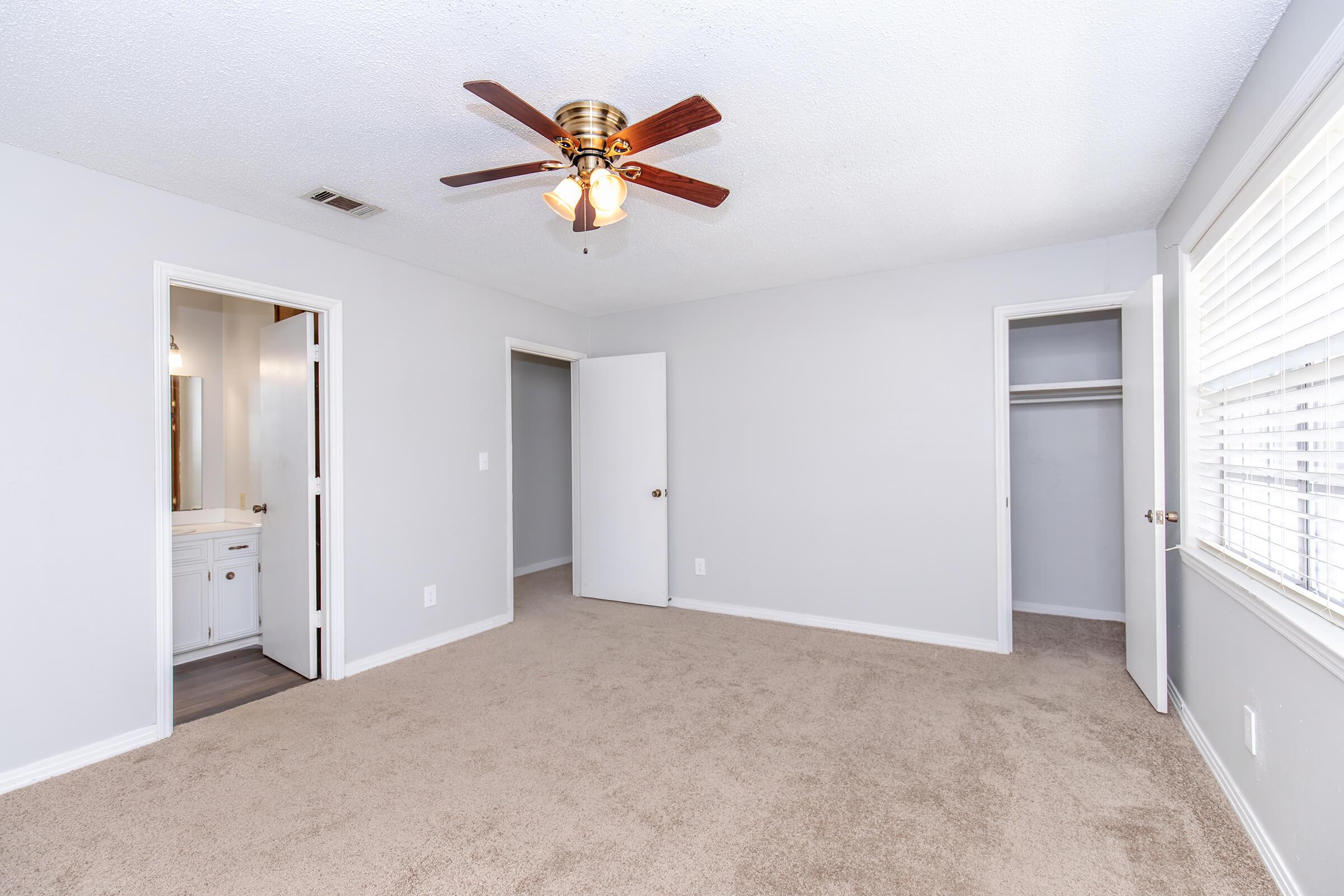 A modern, empty bedroom featuring light gray walls and soft beige carpeting. There is a ceiling fan with a light fixture, and two doors leading to an adjacent room and a closet. A window with blinds provides natural light, adding to the spacious feel of the room.