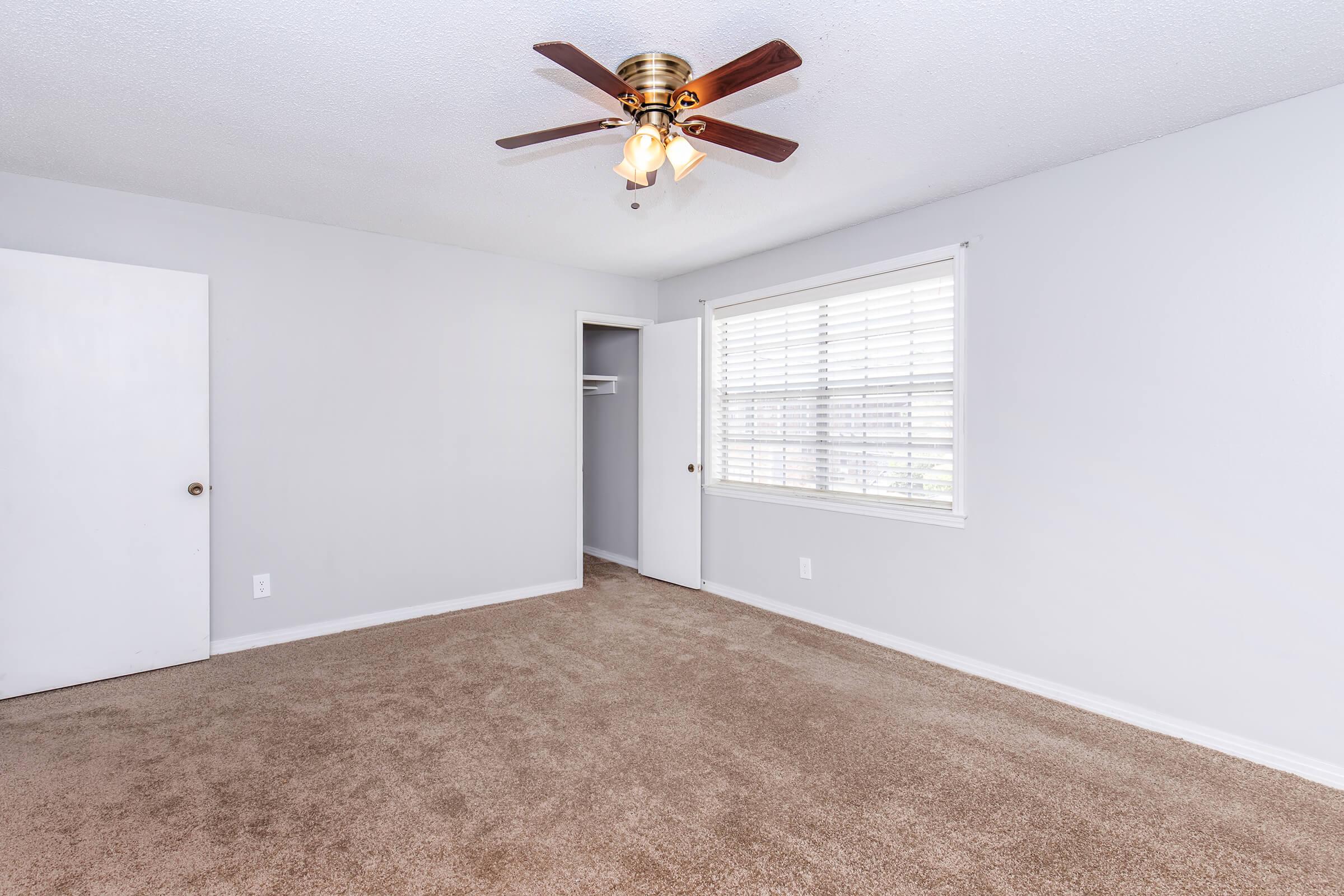 Empty room featuring light gray walls and carpeted flooring. A ceiling fan with light is mounted in the center. One wall has a window with blinds, allowing natural light to enter, while another wall includes a closed door leading to a closet space. The overall ambiance is clean and spacious.