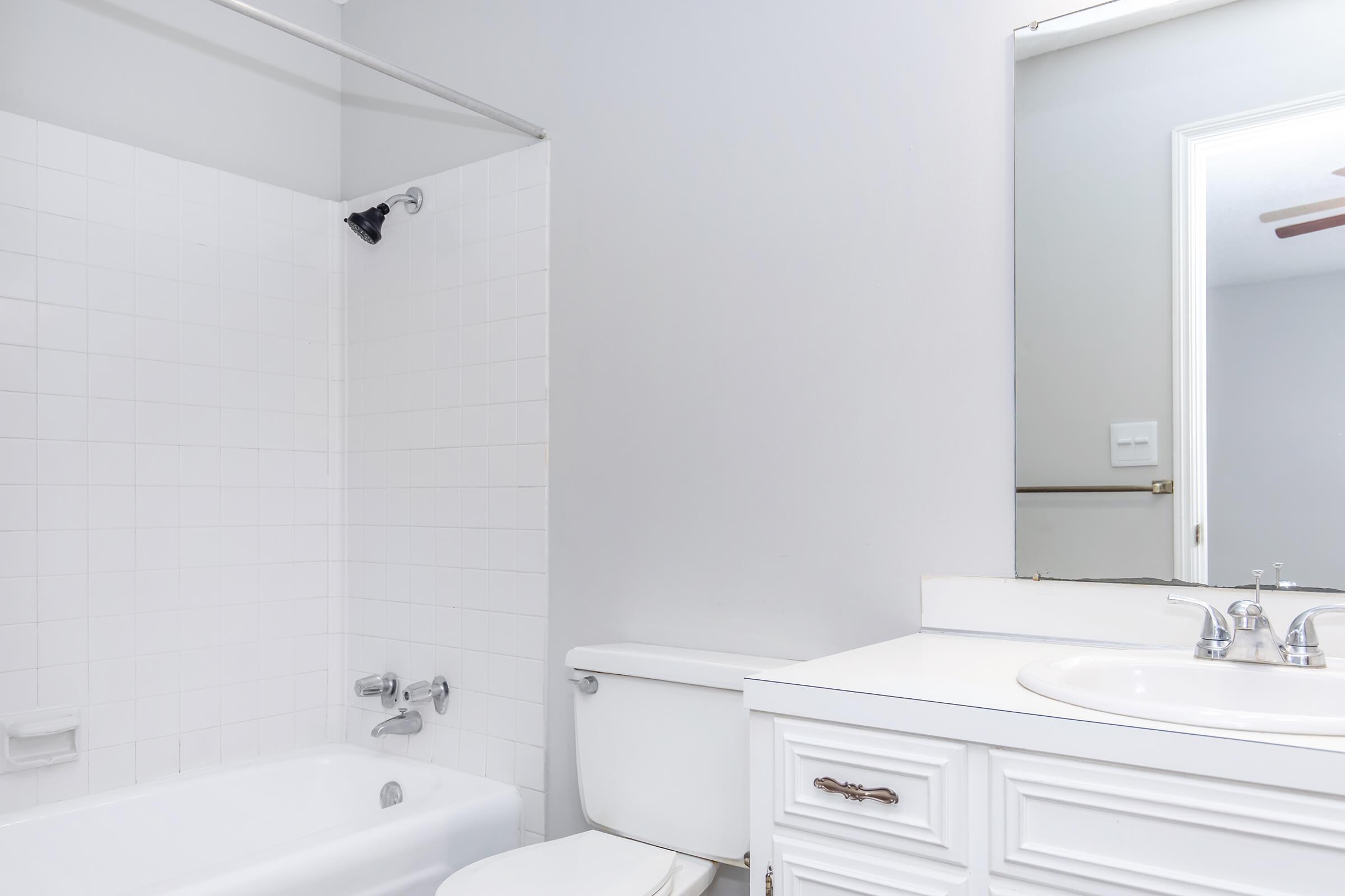A clean, modern bathroom featuring a white bathtub with a showerhead, a white toilet, and a white vanity with a sink. The walls are painted in a light color, and there is a mirror above the sink, contributing to a bright and airy atmosphere.