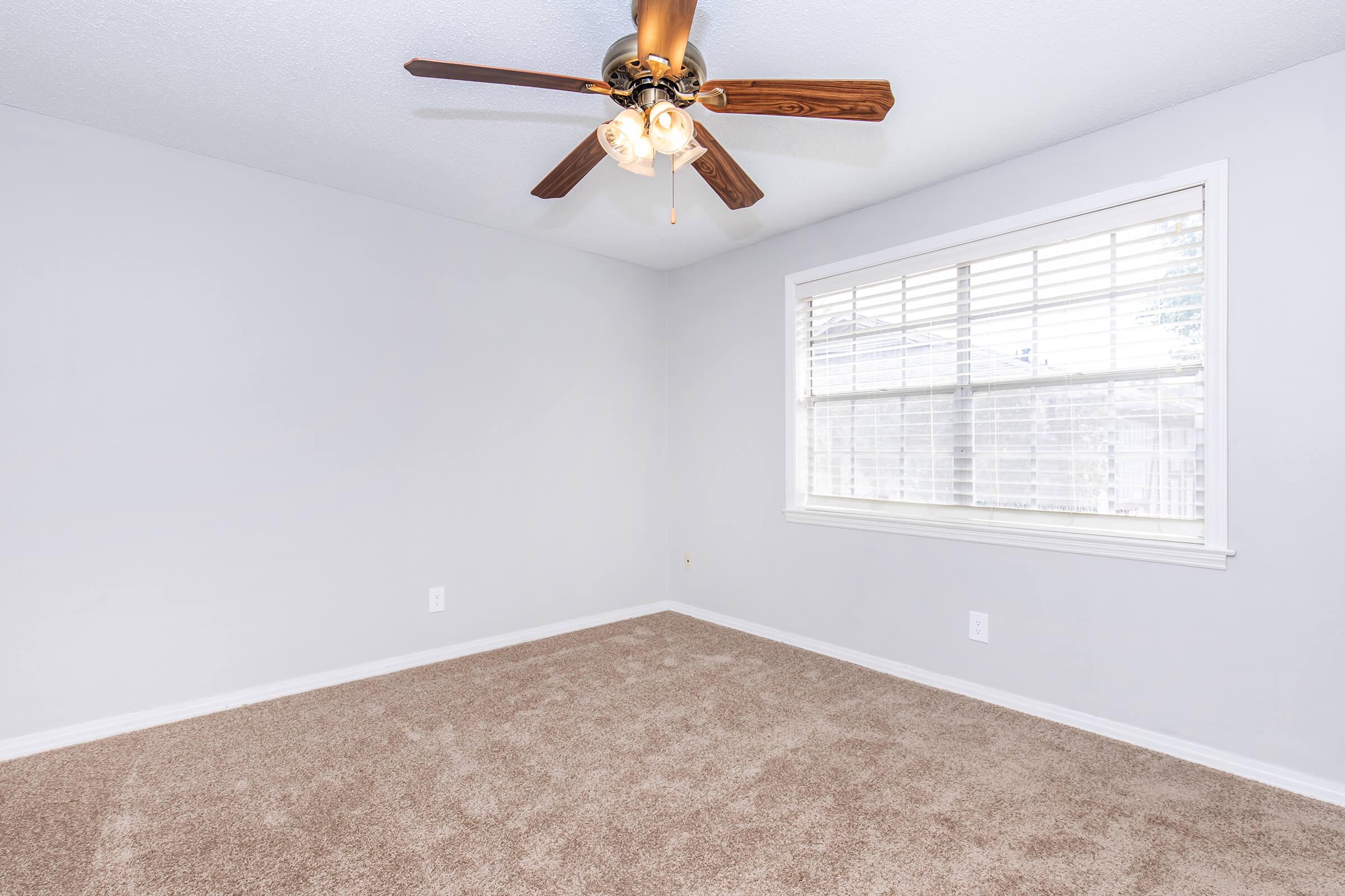 An empty room with light gray walls and beige carpet. A ceiling fan with wooden blades and a light fixture hangs in the center. A large window with white blinds lets in natural light, and there are no furnishings or decorations in the space.