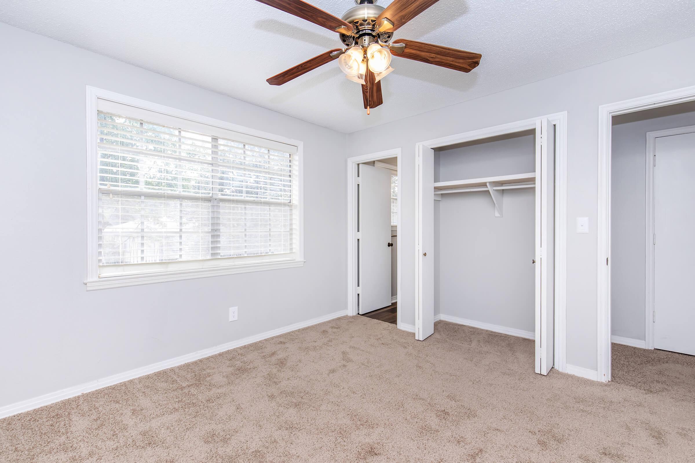 A spacious bedroom featuring a ceiling fan with wooden blades, light-colored walls, and plush carpeting. The room includes a large window with blinds, providing natural light, and has a closet on one side. Two doorways lead to adjacent areas, enhancing the open feel of the space.