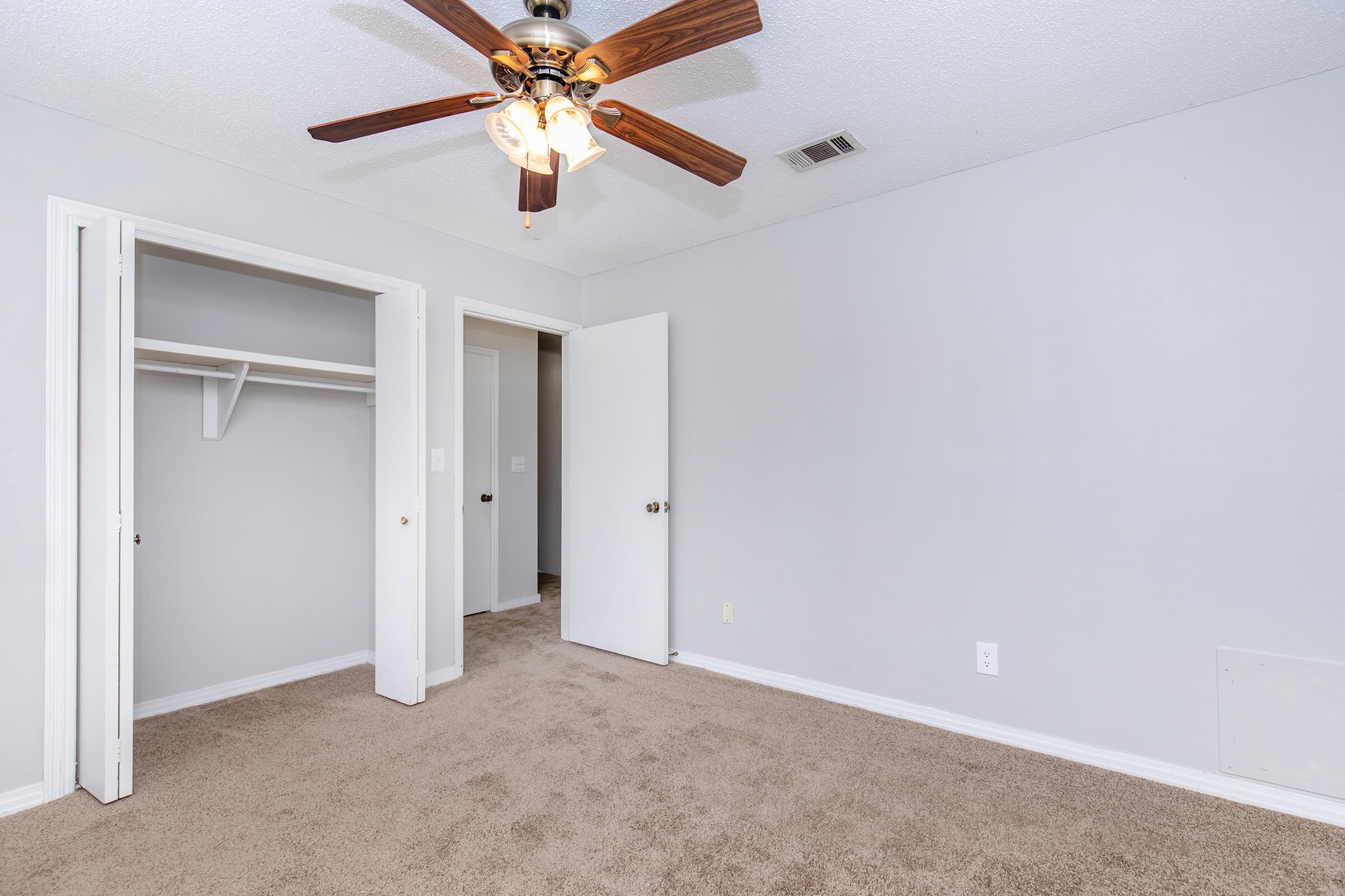 A clean, empty room with light gray walls and beige carpeting. A ceiling fan with three light bulbs hangs from the ceiling. There are two doors: one leading to a closet, and another opening to an adjacent space. Natural light illuminates the area, creating a spacious feel.