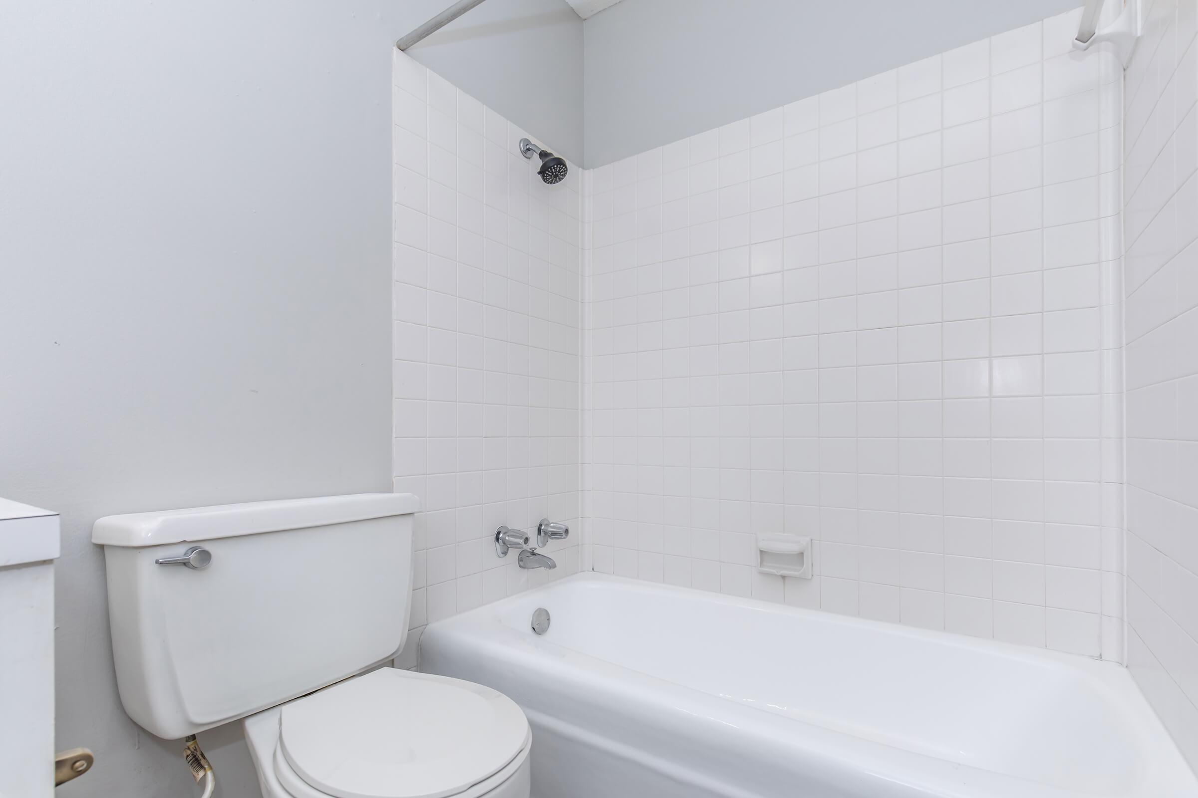 A clean bathroom featuring a white bathtub and a toilet. The walls are tiled with white square tiles, and there is a showerhead above the bathtub. The overall decor is simple and modern, with a neutral color scheme.