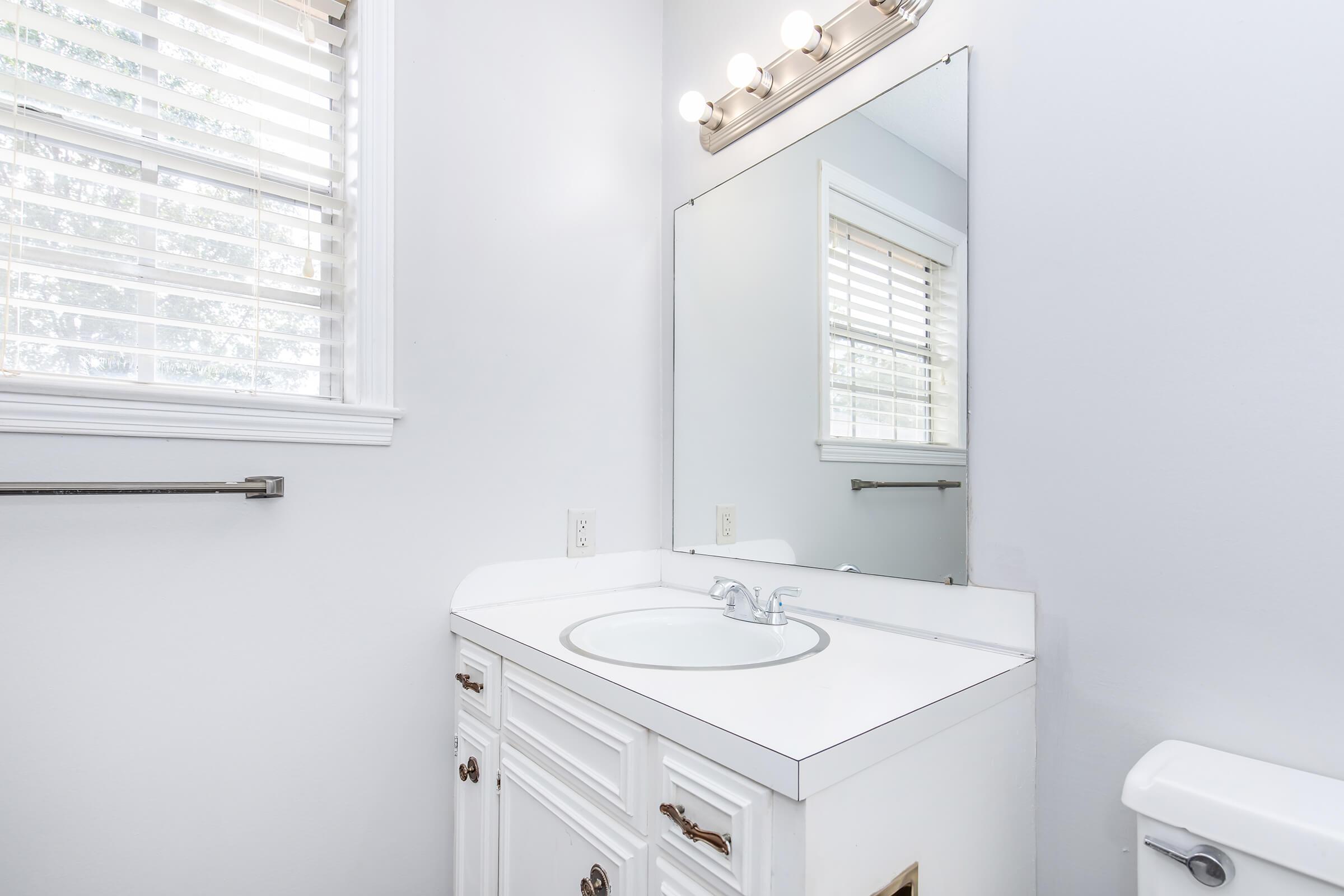 A bright bathroom featuring a white vanity with a circular sink, a large mirror above it, and a light fixture with multiple bulbs. The walls are painted in a light color, and there are white window blinds providing natural light. A toilet is visible to the right, and a towel rack is mounted on the wall.