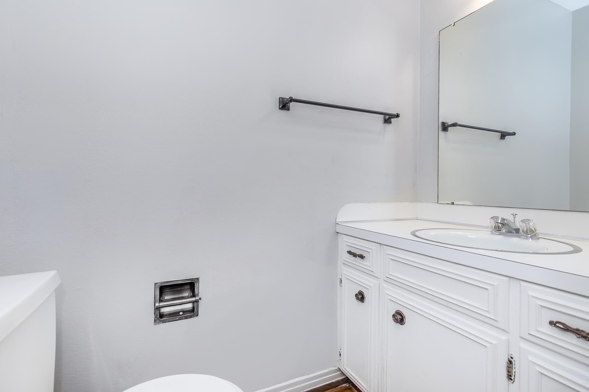 A clean and simple bathroom featuring a white vanity with a round sink, a large mirror above the sink, a toilet, and a towel rack on the wall. The walls are painted light gray, and the flooring is wooden. The overall aesthetic is minimalist and tidy.