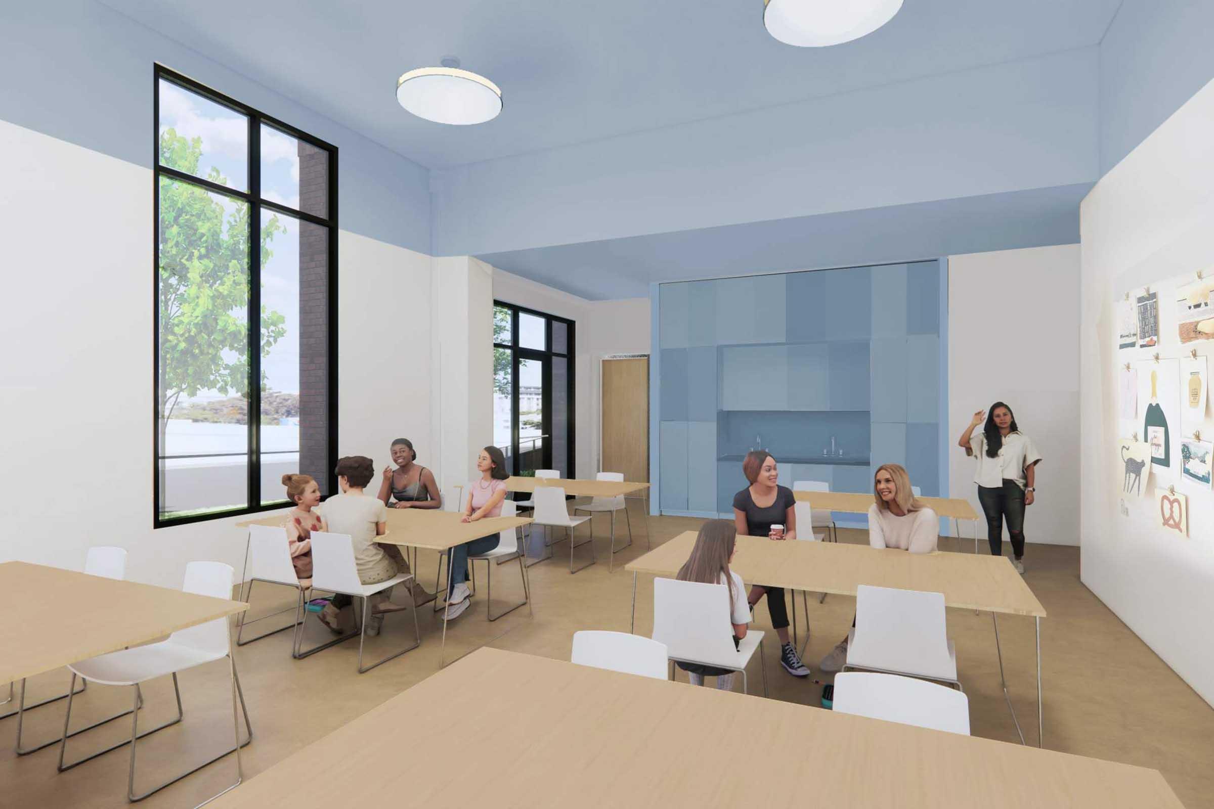 A bright, modern communal space featuring several tables with chairs. Groups of people are engaged in conversation or activities. Large windows let in natural light, and there are trees visible outside. One woman stands in the background, while others sit and socialize around tables.