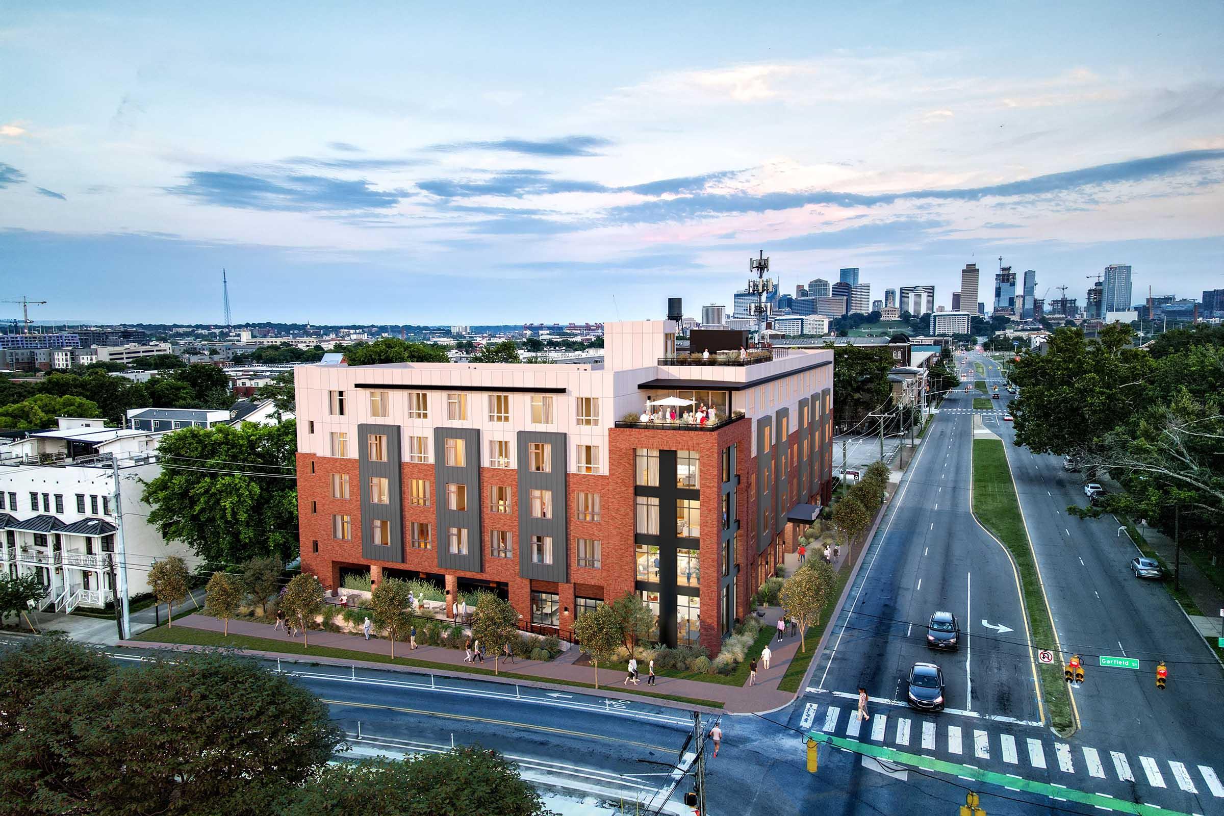 A modern, multi-story brick building with large windows, surrounded by greenery and trees, situated along a road. The skyline of a city with tall buildings is visible in the background under a cloudy sky, suggesting a vibrant urban environment.