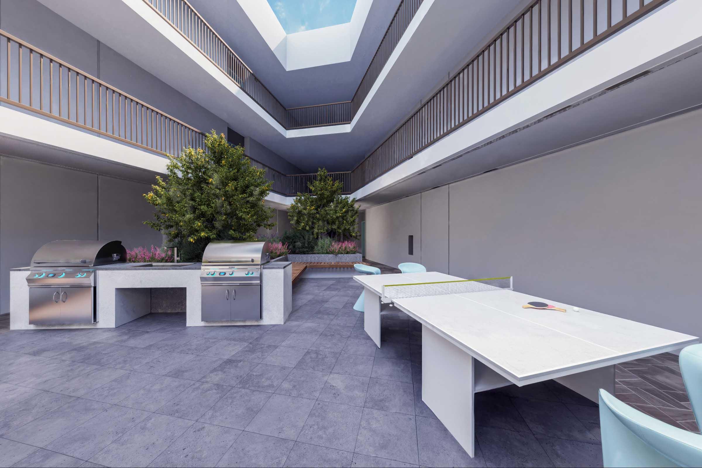 A modern outdoor space featuring a patio with two grilling stations, surrounded by greenery and flowering plants. The area has sleek, minimalist furniture, including a large table and chairs, under an open sky with a glass ceiling above, creating a light and airy atmosphere.