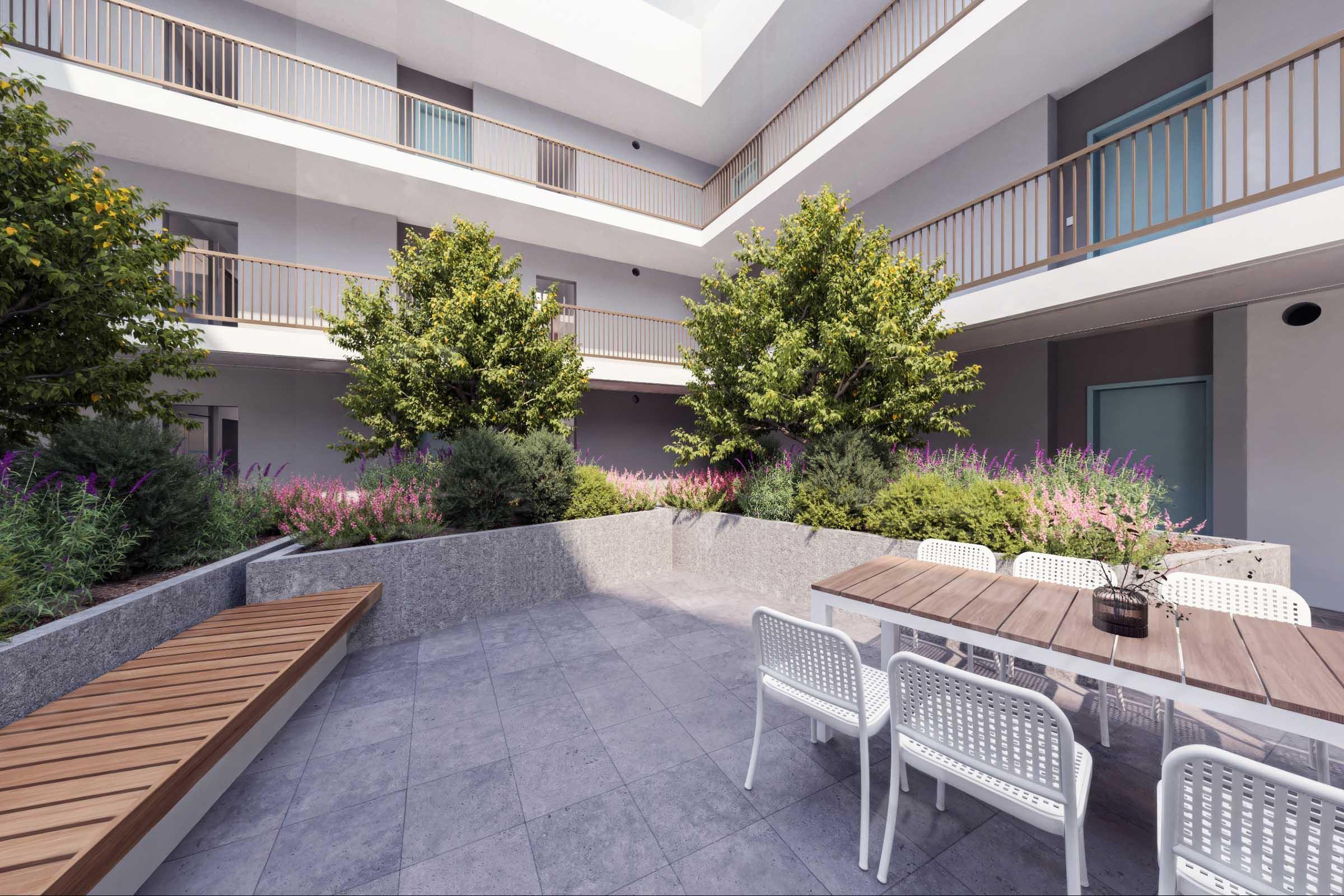 A modern courtyard with two trees and colorful flowering plants. There is a rectangular stone planter and a wooden bench along one side. A white outdoor dining table with chairs is set in the center, surrounded by a contemporary building featuring balconies. Soft natural lighting brightens the space.
