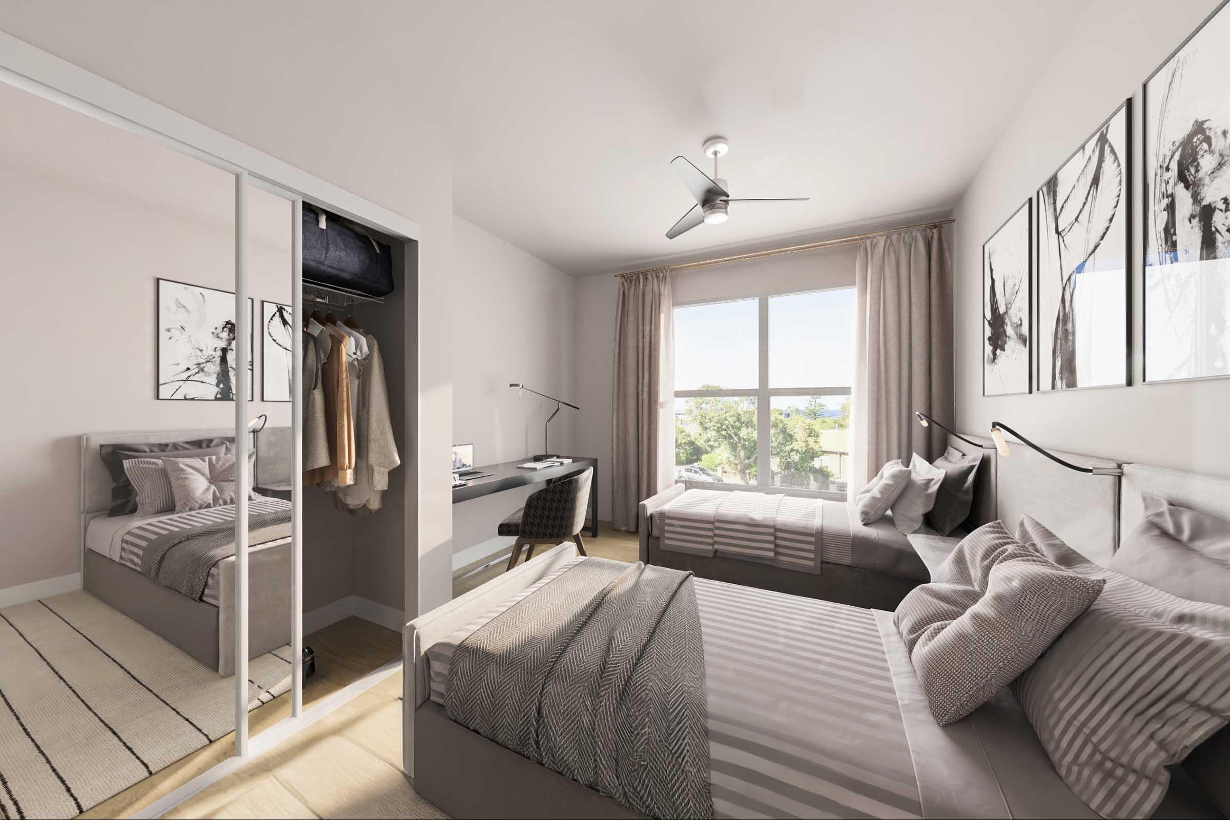 A modern bedroom featuring two single beds with gray bedding, a large window letting in natural light, and sheer curtains. A desk with a reading lamp is positioned near the window, and a mirrored closet reflects part of the room. Two framed artworks adorn the walls.