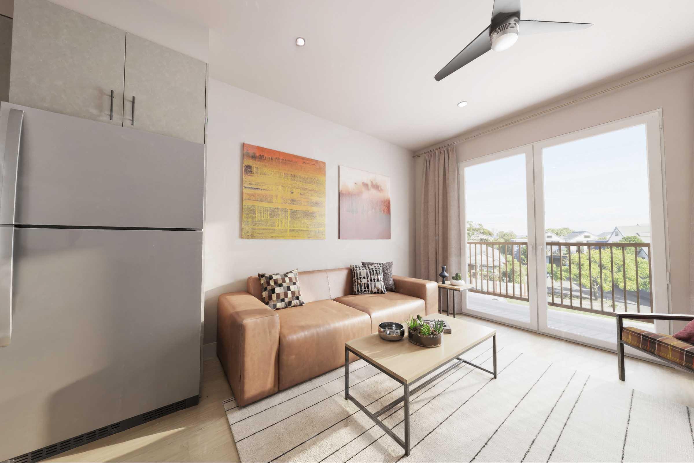 Interior view of a modern living room featuring a brown leather sofa, a glass coffee table, and a small indoor plant. Two abstract wall art pieces are displayed above the sofa. A refrigerator is visible in the corner, and large windows provide natural light and a view of the outdoors.