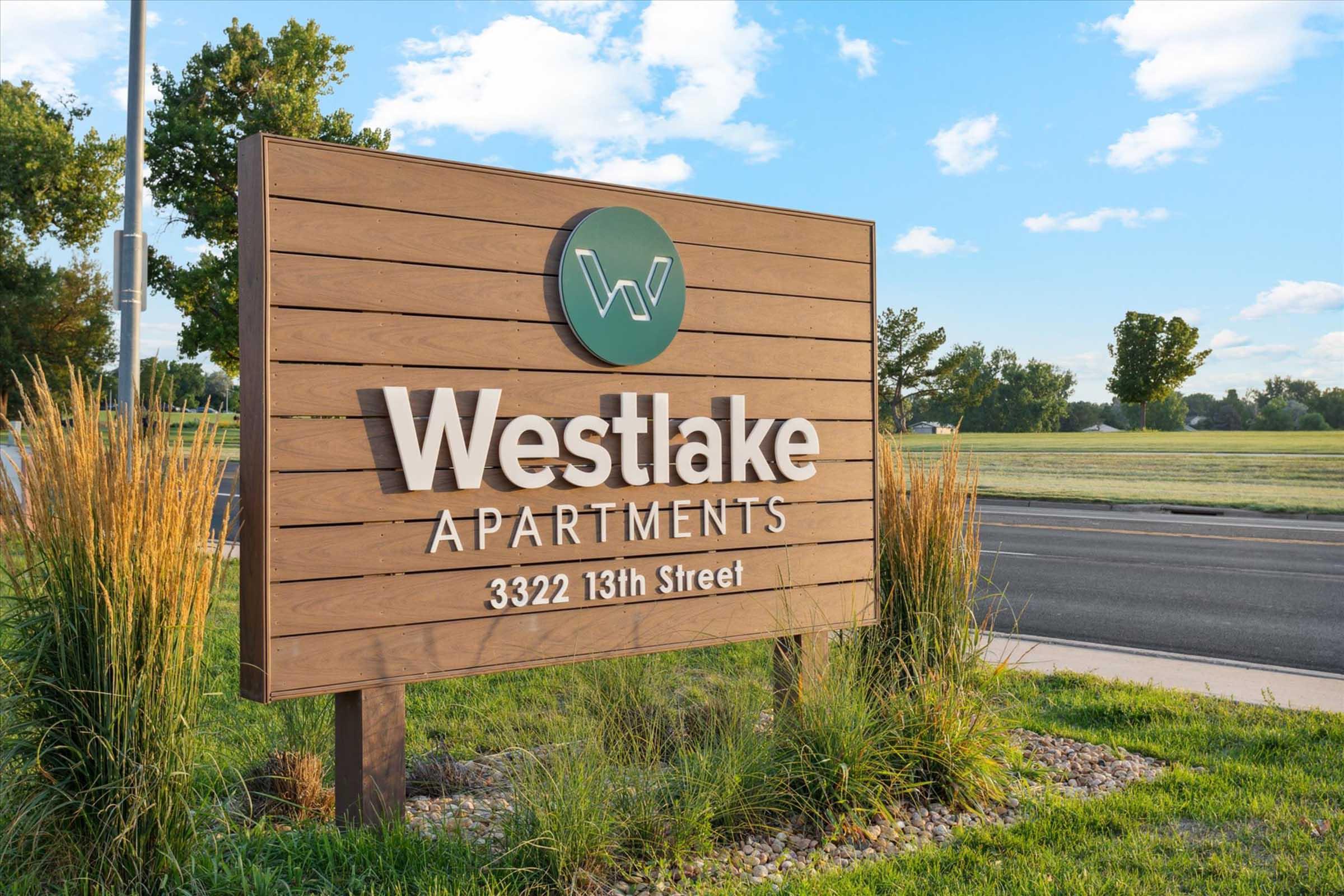 Wooden sign for Westlake Apartments, displaying the name prominently in white letters against a natural backdrop of grass and trees. The sign features a circular logo in green with a stylized 'W'. Located at 3322 13th Street, the scene is set against a clear blue sky.