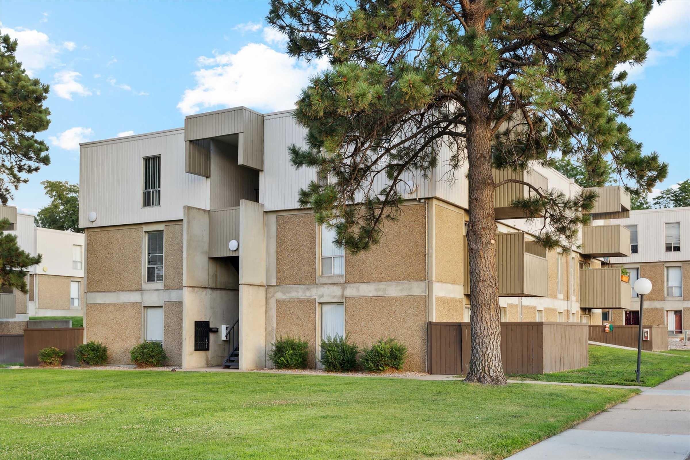 Low-rise apartment building with a mix of textured beige and gray siding. Surrounded by green grass and a few shrubs, a large pine tree is in the foreground. The building features multiple balconies and large windows, with outdoor lighting fixtures visible. Clear blue sky in the background.
