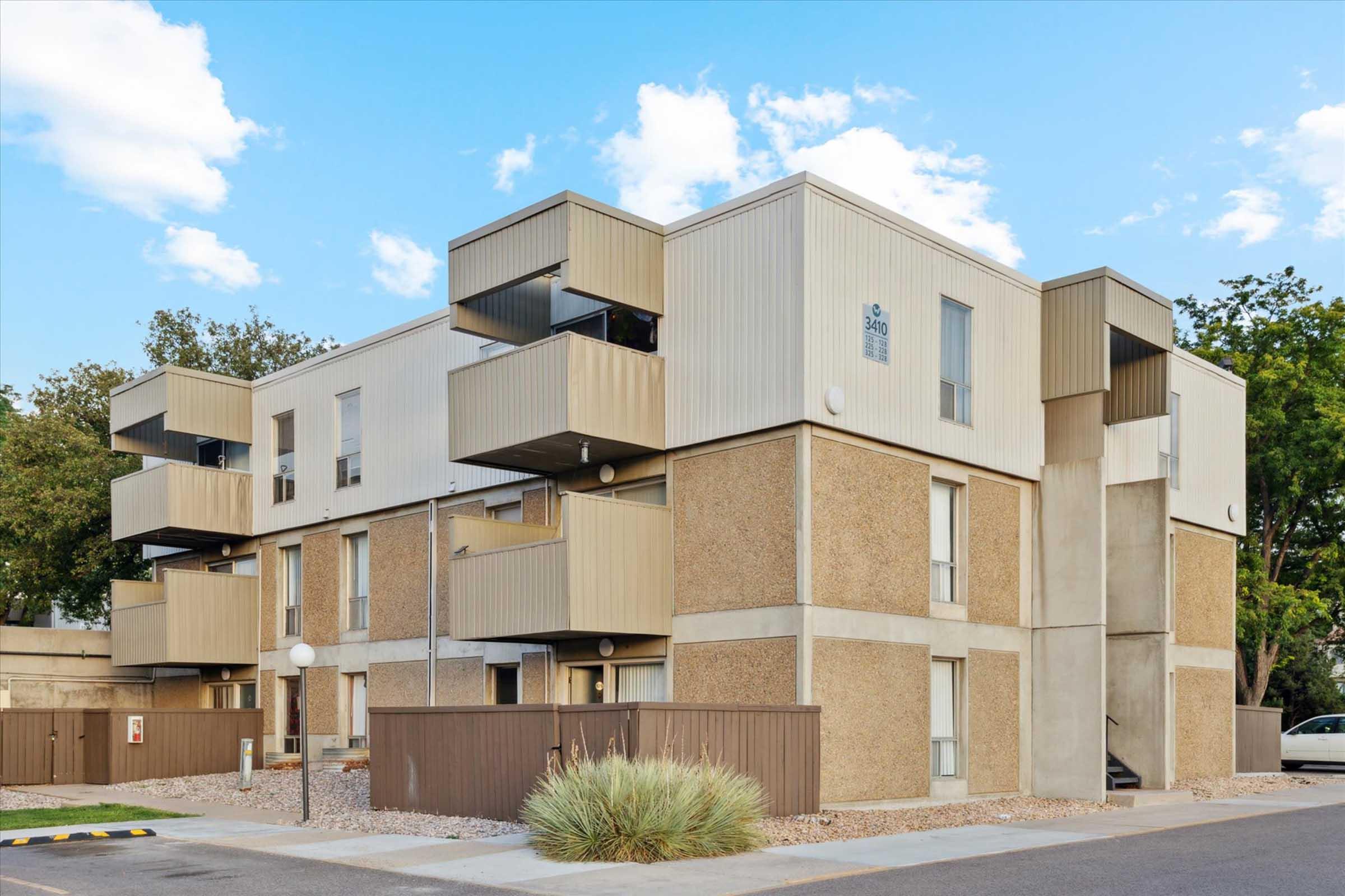 A modern, multi-story apartment building with a tan exterior, featuring several balconies. The structure is surrounded by a landscaped area with low shrubs and a clear blue sky in the background. The driveway has parking spaces and a sign indicating the building number.