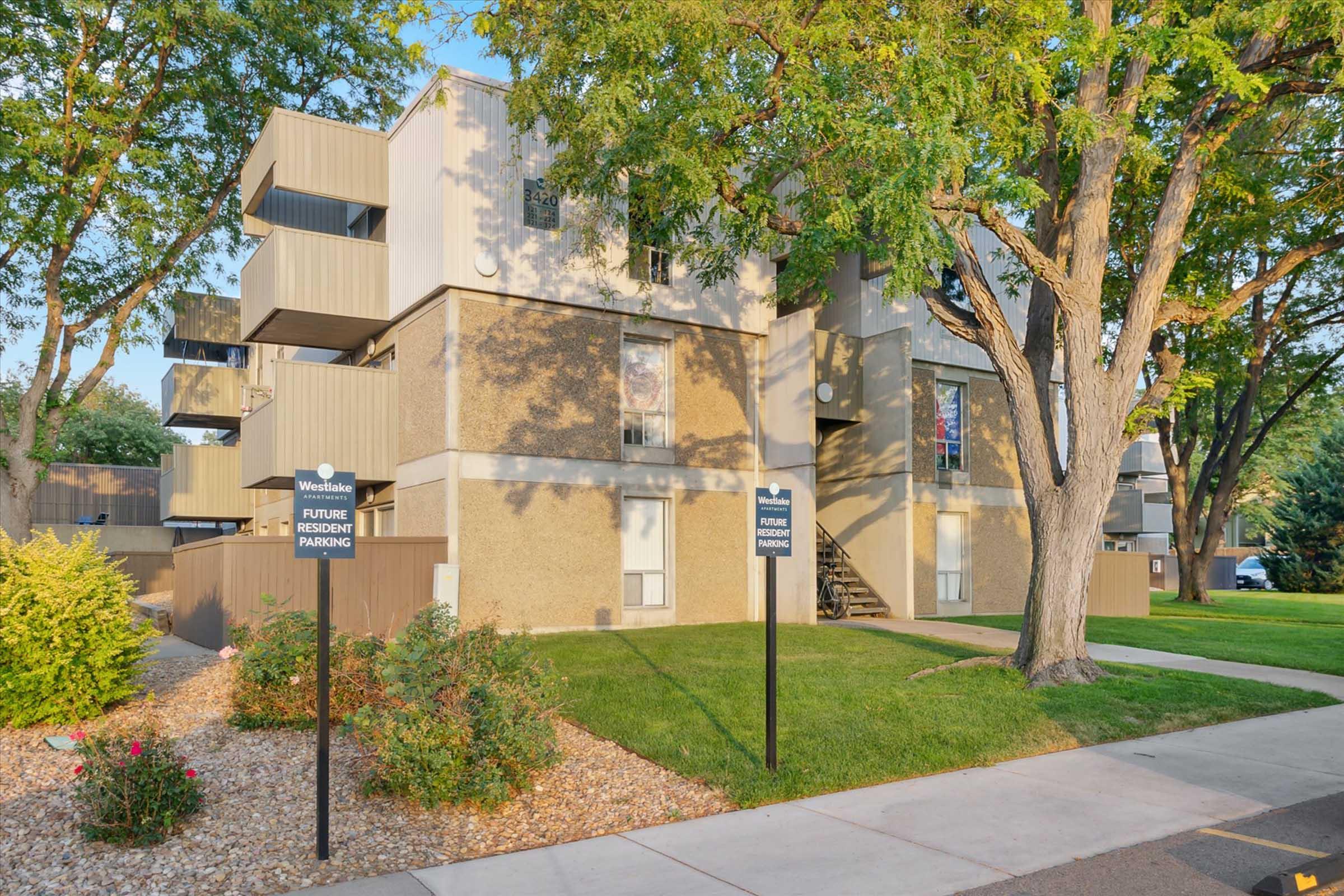 A residential building with three levels, featuring balconies and surrounded by greenery. Signs indicate "Future Resident Parking" and "Visitor Parking." Well-maintained landscaping includes shrubs and small flowers, with a sidewalk leading to the entrance.