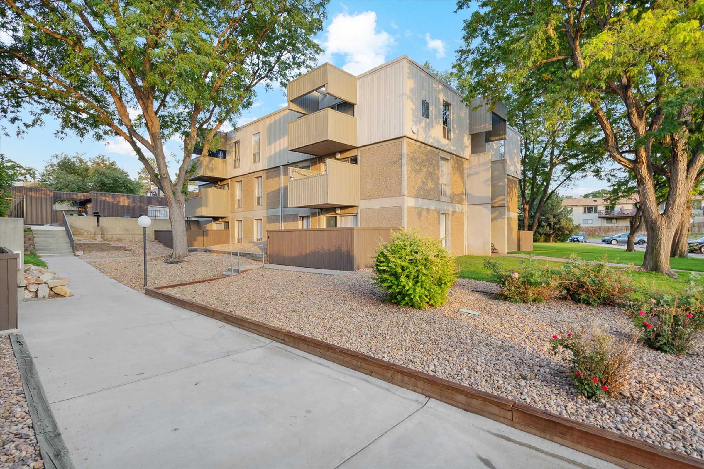 A view of a modern apartment complex surrounded by trees and landscaped areas. The building features multiple balconies, a light post, and gravel pathways. Grass and plants complement the outdoor scene, contributing to a welcoming residential environment in a suburban setting.