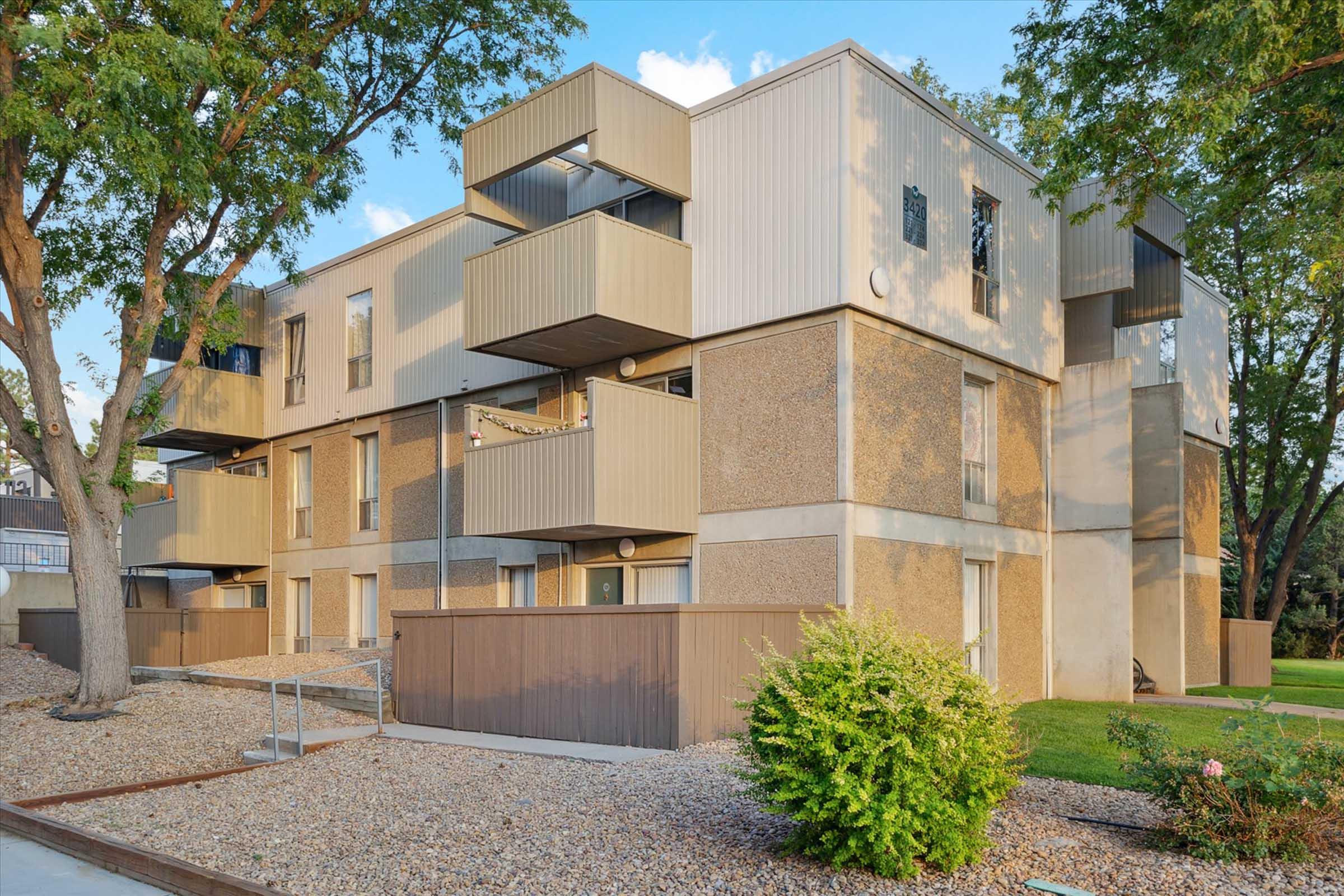 A modern apartment building featuring multiple levels, with a mix of textured and smooth exterior walls. Balconies are visible on the upper floors, surrounded by landscaped greenery and a rocky pathway. Mature trees provide shade in the area.