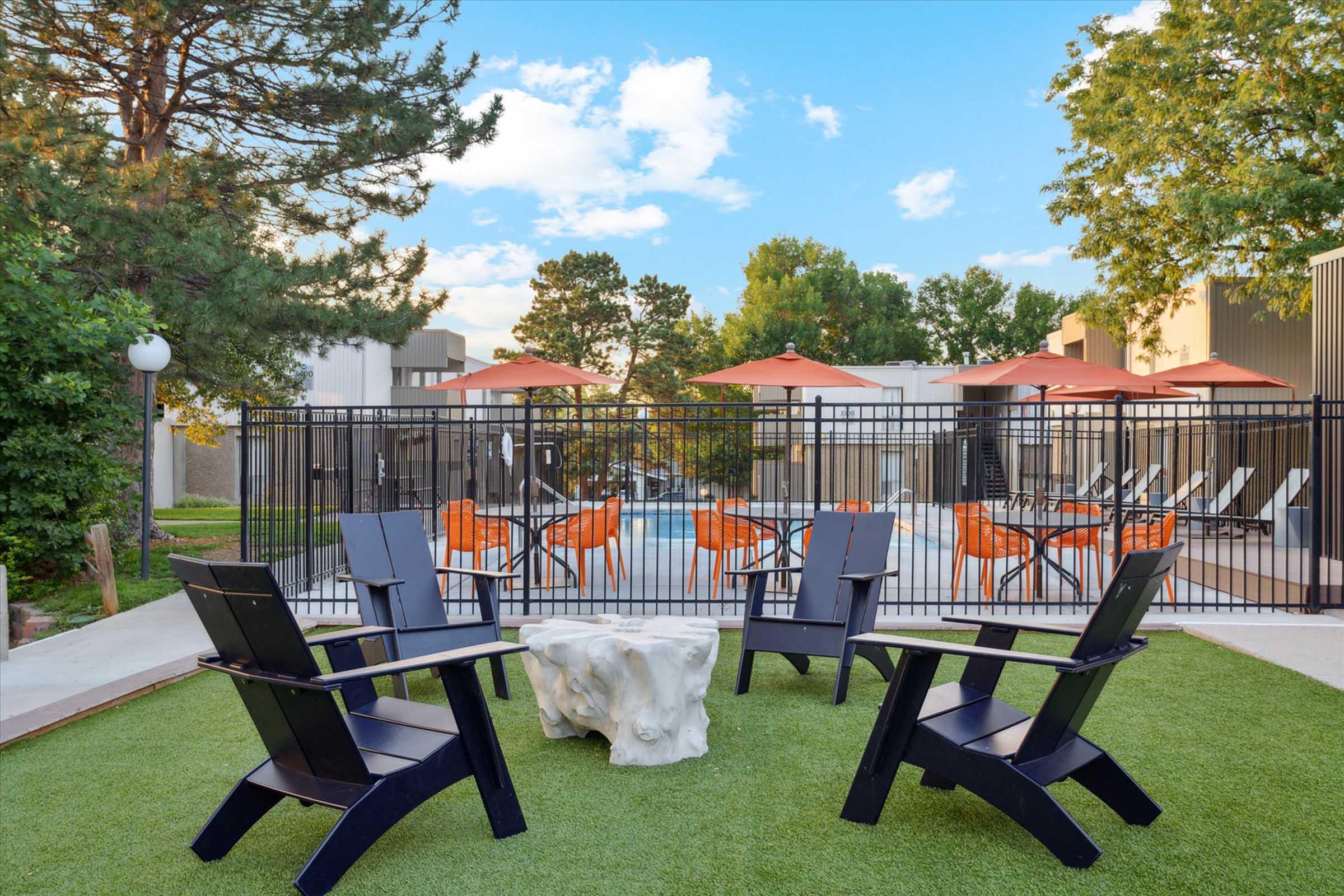A cozy outdoor lounge area featuring two black Adirondack chairs and a unique, organic-shaped table on green grass. In the background, there are orange umbrellas over seating by a pool, surrounded by modern buildings and trees under a blue sky with fluffy clouds.