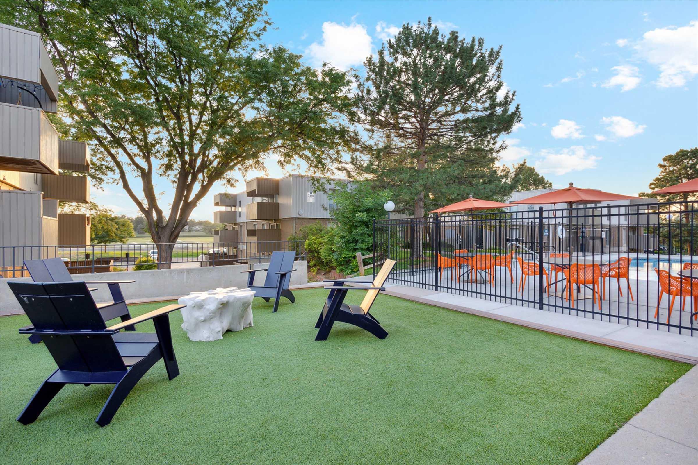 Cozy outdoor space featuring green artificial grass, stylish black and wooden chairs arranged around a stone table. In the background, there are orange umbrellas over a dining area and modern apartment buildings framed by lush trees and a clear blue sky.