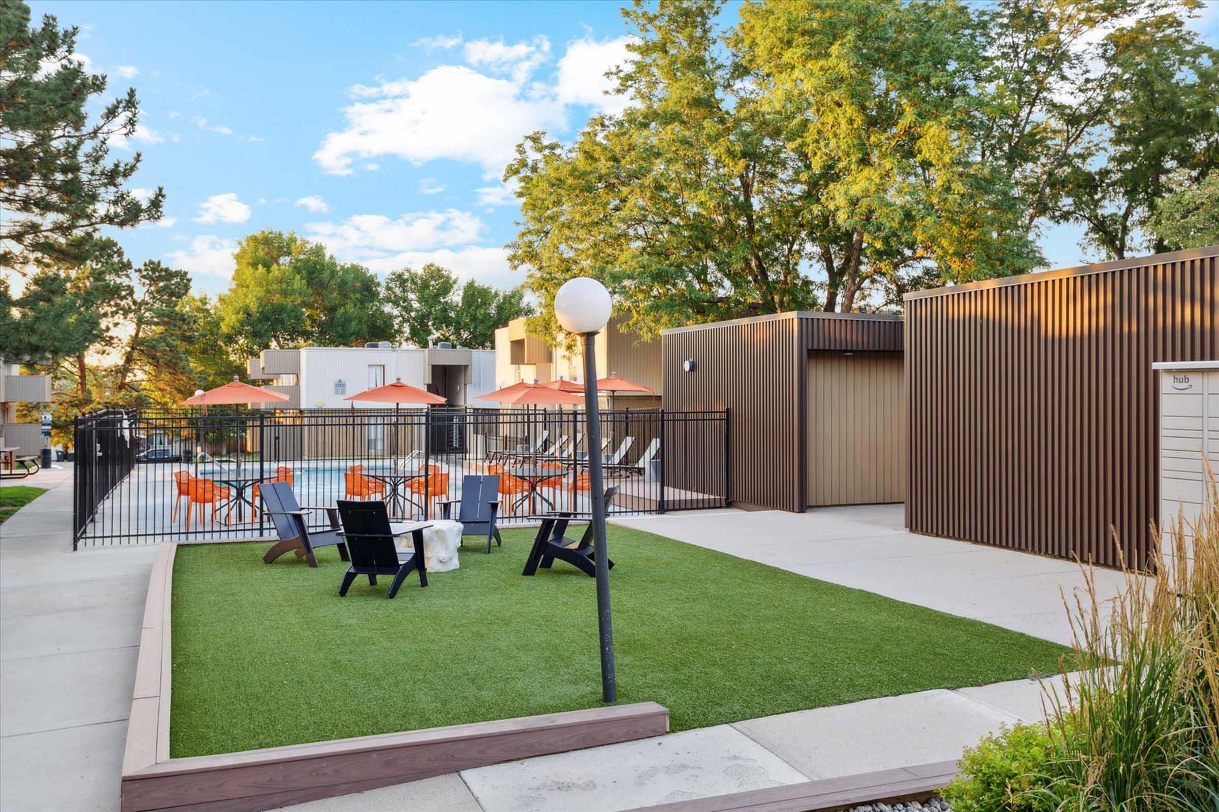 A well-maintained outdoor area featuring green grass, lounge chairs, and orange umbrellas around a pool. Surrounding trees provide shade, and modern buildings are visible in the background. The space is fenced for privacy, creating a relaxing atmosphere for residents.