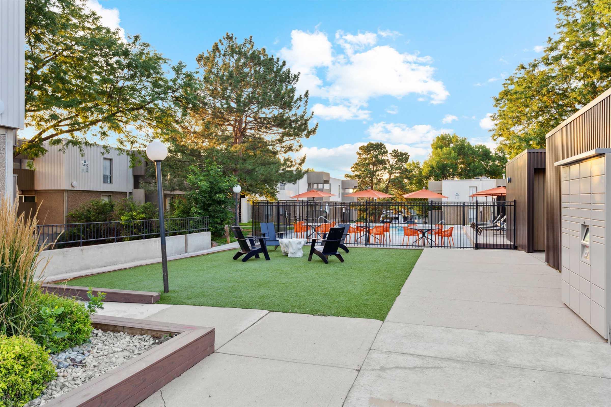A well-maintained outdoor space featuring a small green lawn, black lounge chairs, and a paved walkway. In the background, there are orange patio chairs around a pool area, surrounded by trees and buildings, under a blue sky with fluffy clouds.