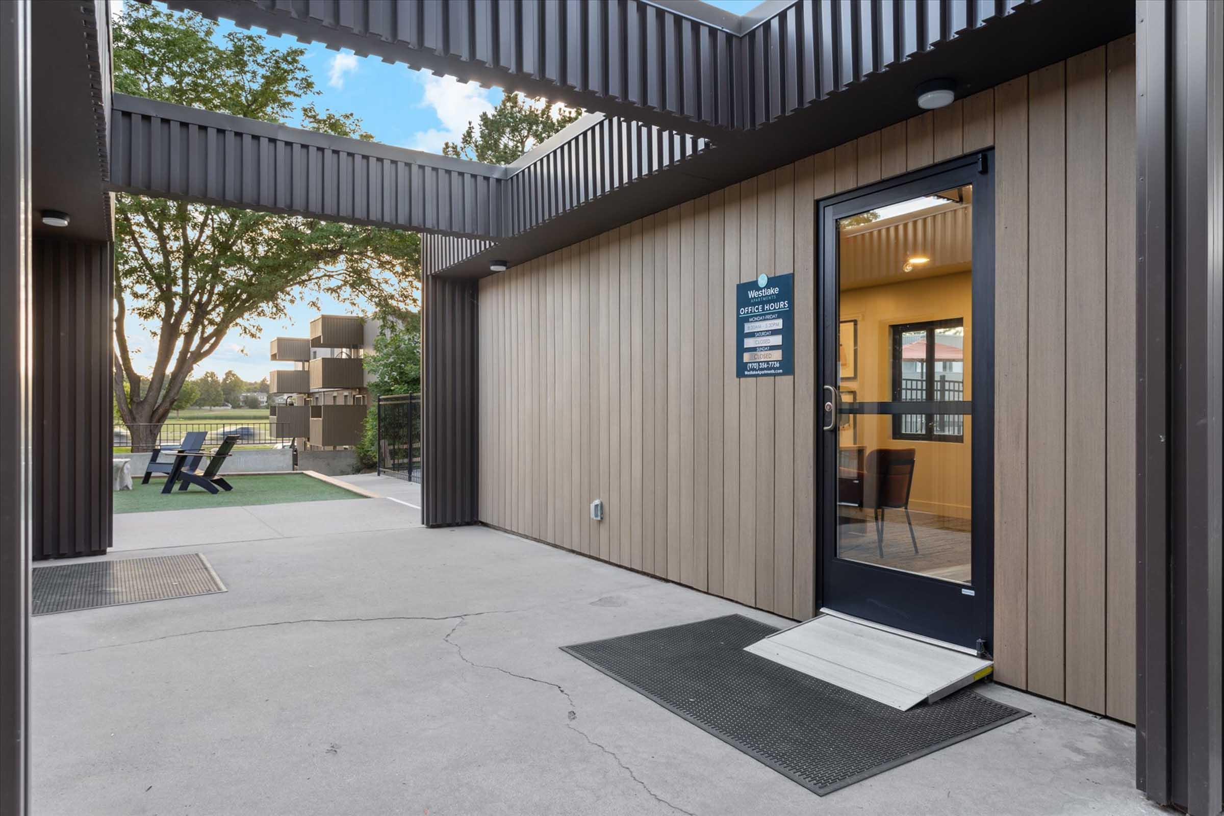 A modern building entrance featuring a black door with glass panels, surrounded by a wooden facade and metal accents. The area outside has a concrete floor, greenery visible in the background, and seating nearby. Signs are displayed next to the door, indicating the building's purpose.