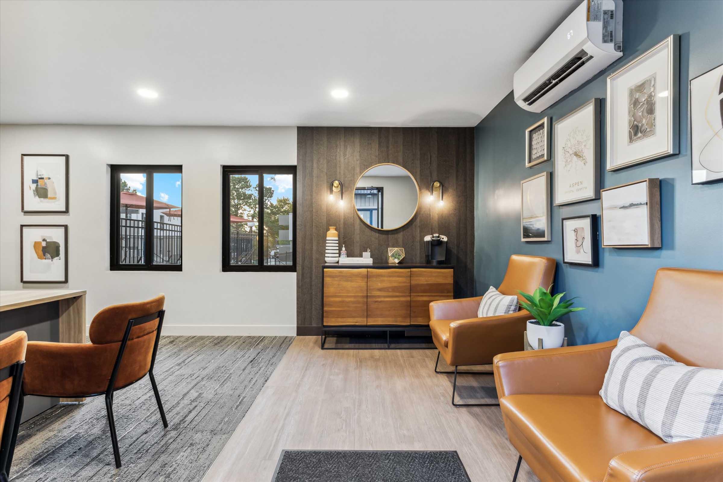 A modern waiting area featuring two brown leather chairs, a wooden side table with decorative items, a large round mirror on the wall, framed artwork, and windows allowing natural light. The room has a stylish gray and blue color scheme with warm lighting.