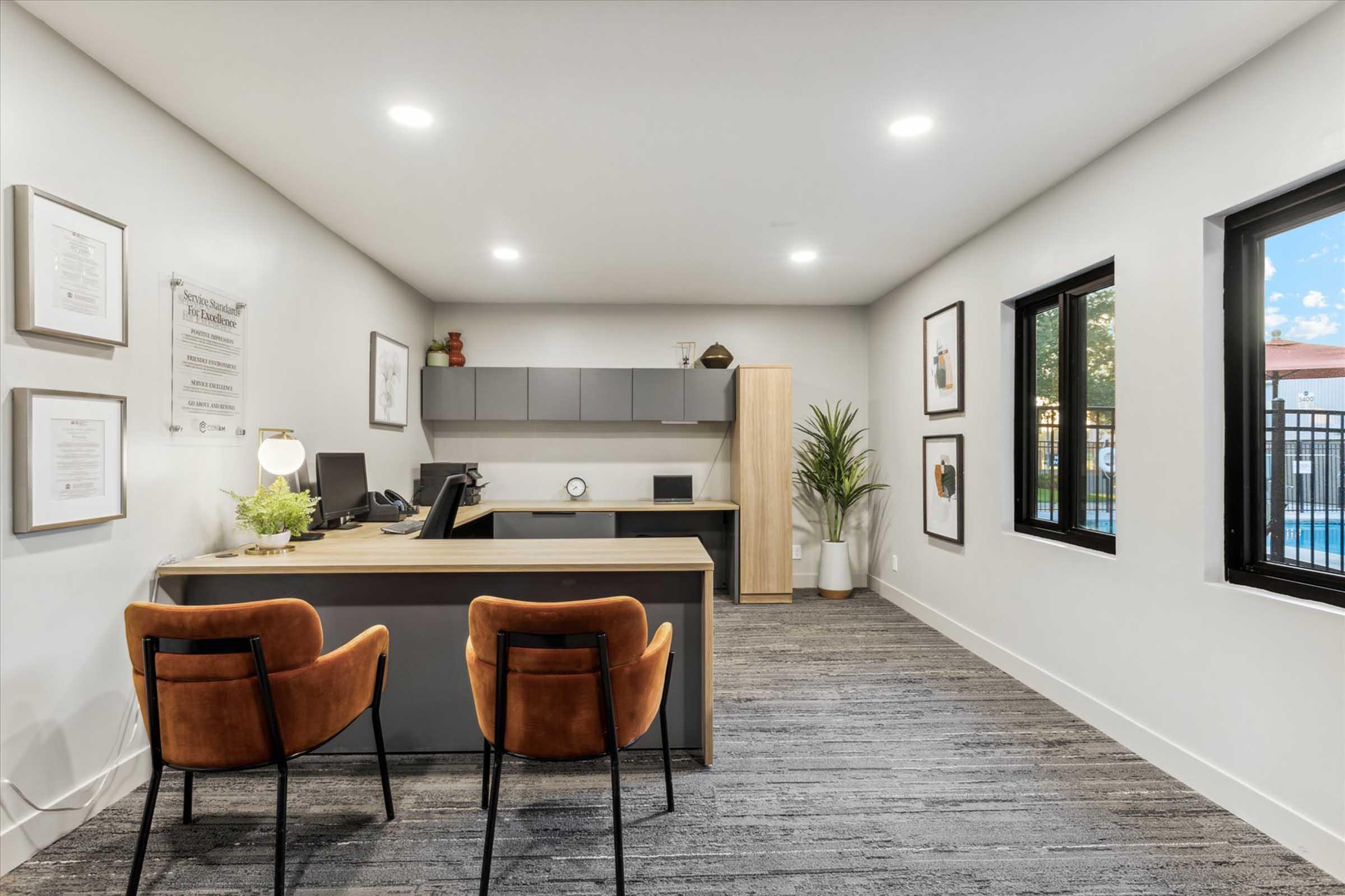 Modern office interior featuring a sleek desk with a computer, two stylish chairs, framed artwork on the walls, and a large potted plant. Large windows showcase outdoor views, and the space has a light color palette with warm lighting, creating a welcoming atmosphere.