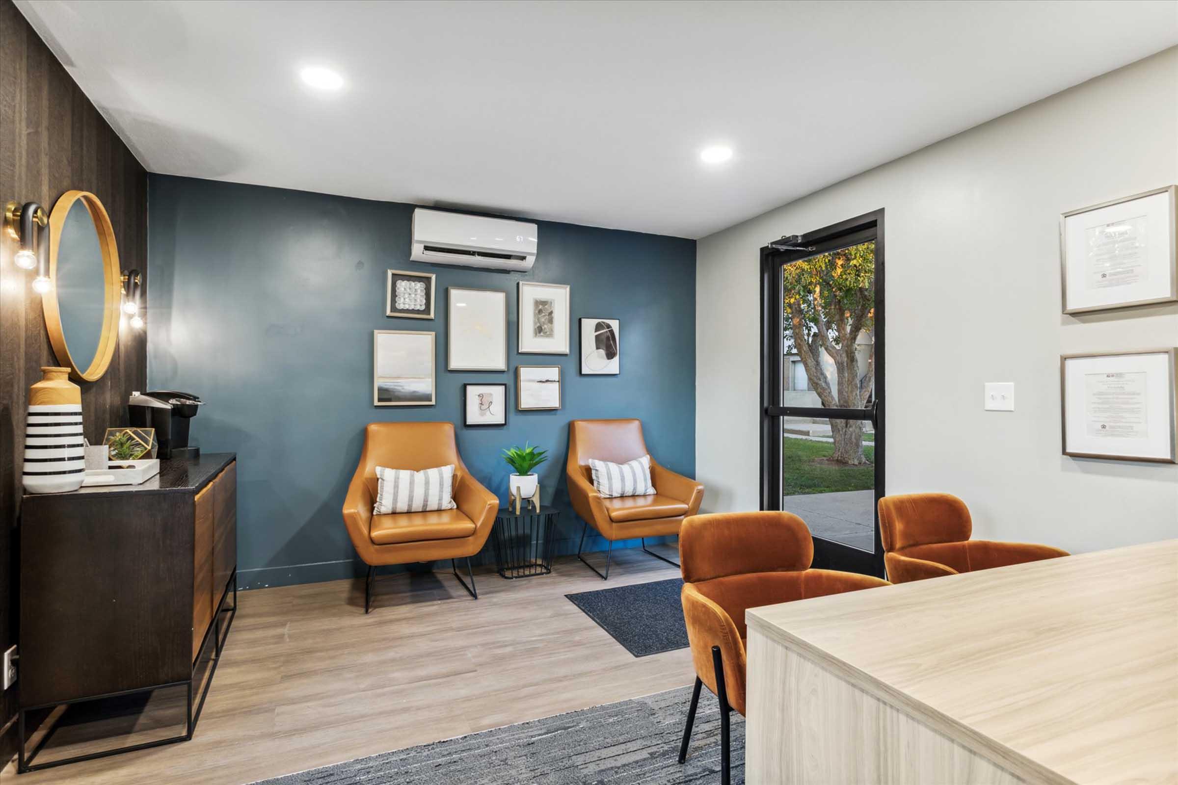 A modern interior of a cozy office space featuring two brown armchairs with white cushions, a small round table, a black cabinet, and wall décor including framed art. Large windows allow natural light, and there is a potted plant adding a touch of greenery to the ambiance.
