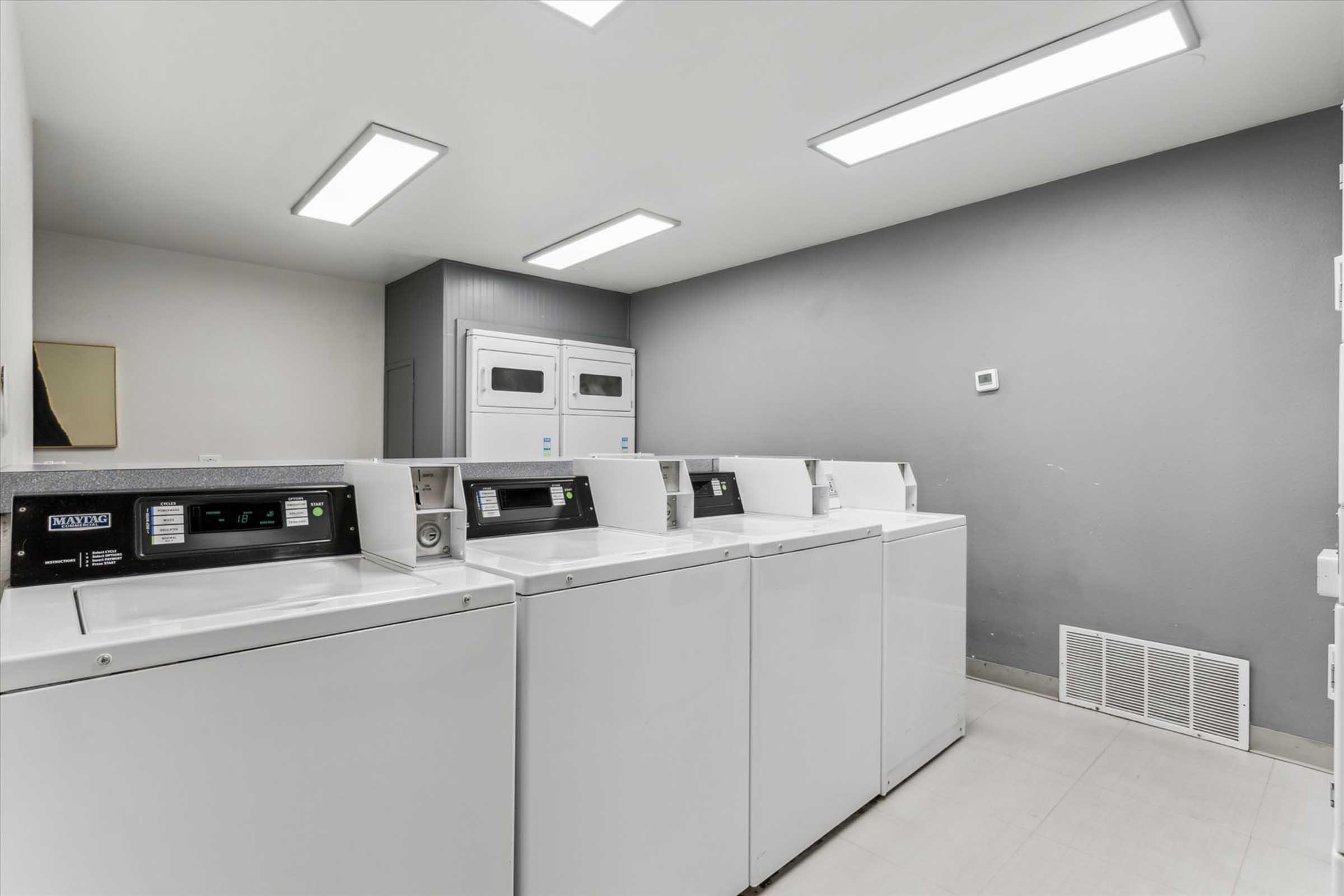 A modern laundry room featuring three row of white washing machines and a dryer at the far end, all against a gray wall. Bright overhead lights illuminate the space, creating a clean and functional appearance.