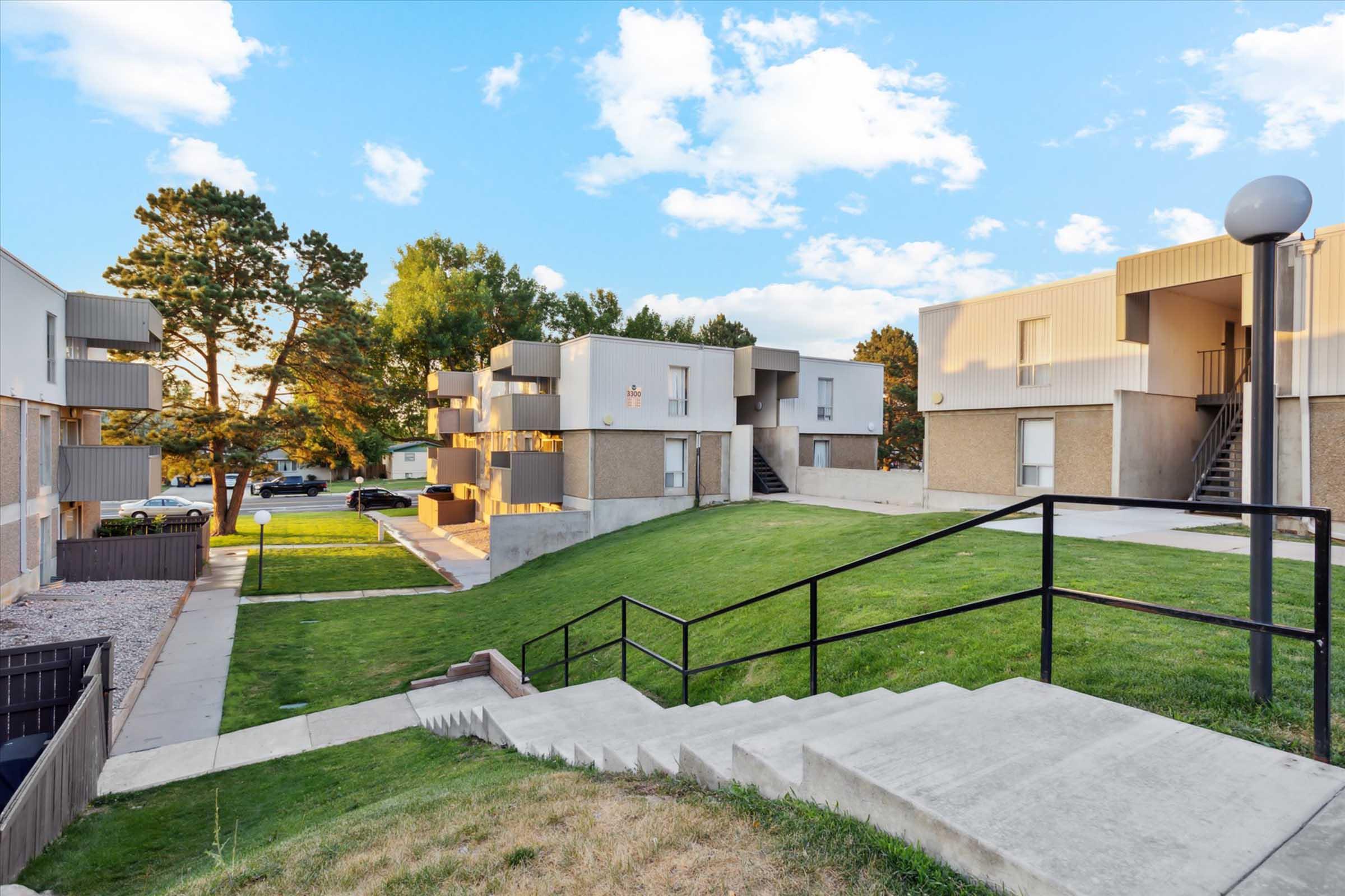 A view of an apartment complex featuring several two-story buildings surrounded by green lawns and trees. A concrete staircase leads down to a pathway, with parked cars visible in the background. The sky is partly cloudy, adding to the peaceful atmosphere of the neighborhood.