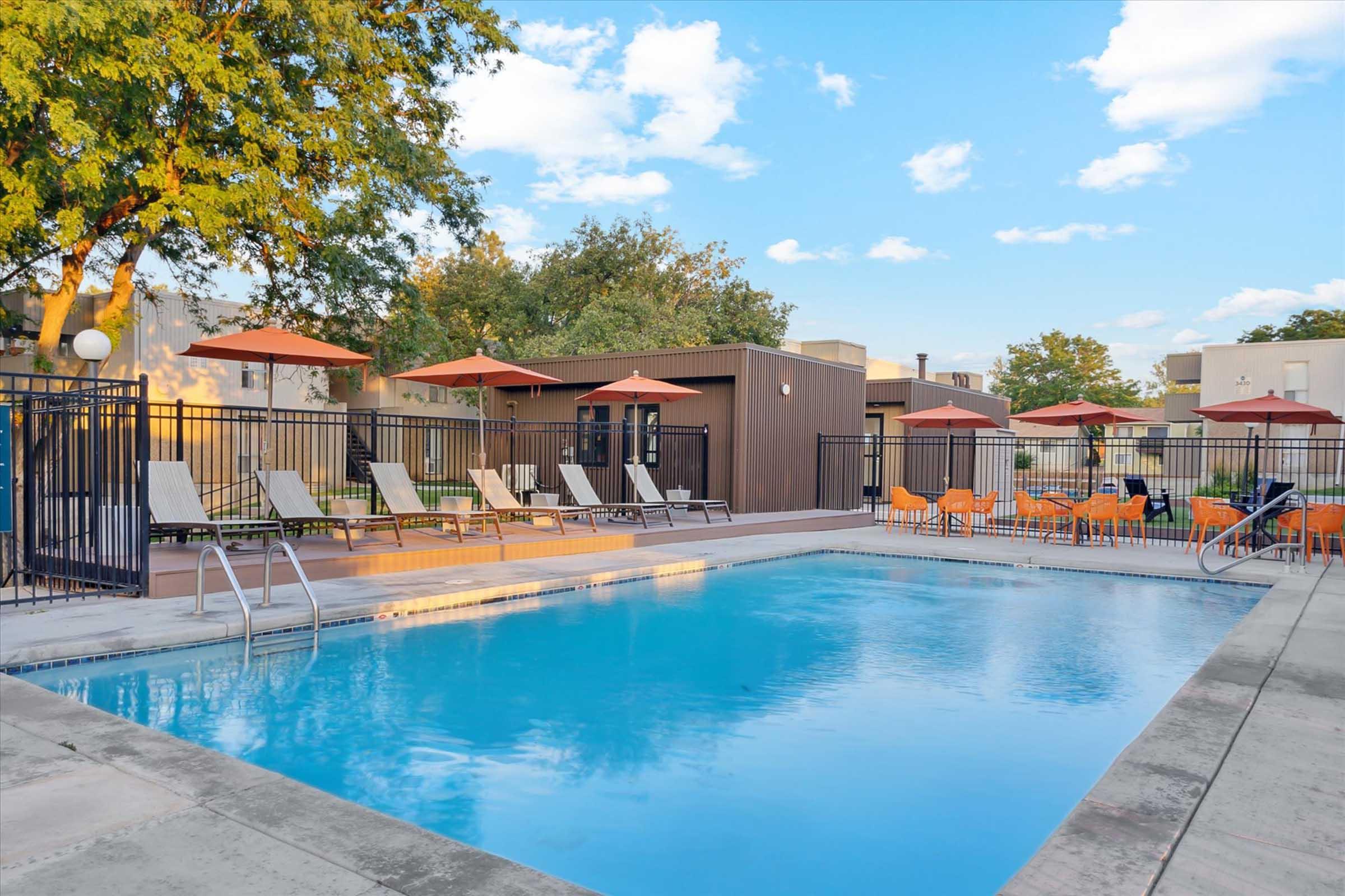 A serene outdoor swimming pool surrounded by lounge chairs and umbrellas, set against a backdrop of clear blue skies and lush trees. The pool area is enclosed by a black fence, with a modern building in the background, creating a relaxing and inviting atmosphere for leisure.