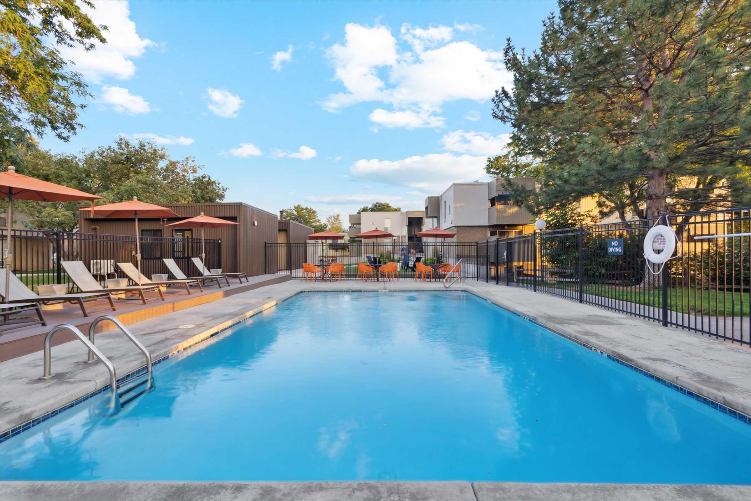 A clear blue swimming pool surrounded by lounge chairs and umbrellas on a sunny day. Green trees are visible in the background, along with modern buildings. The area is well-maintained, creating a relaxing outdoor space for leisure and recreation.