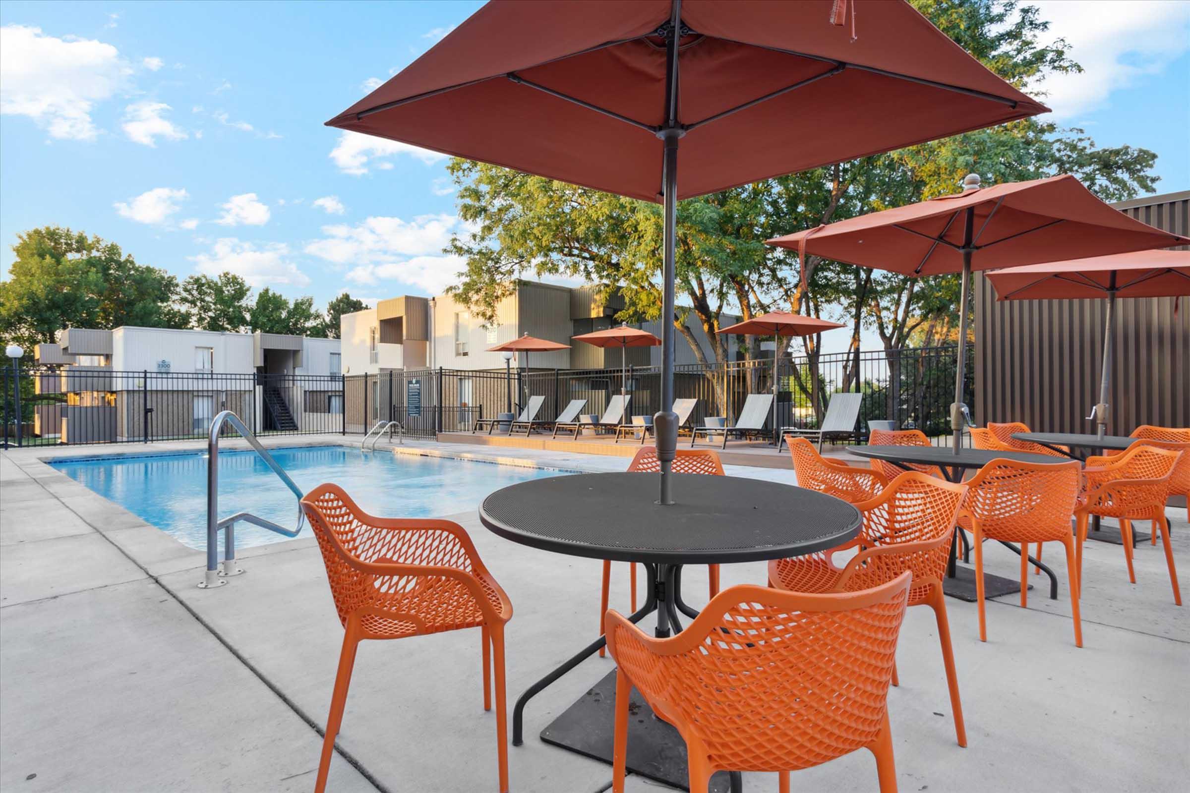 A swimming pool area featuring a clear blue pool, surrounded by orange patio chairs and tables. Several orange umbrellas provide shade, and there are lounge chairs positioned around the pool. In the background, modern buildings and greenery are visible under a partly cloudy sky.
