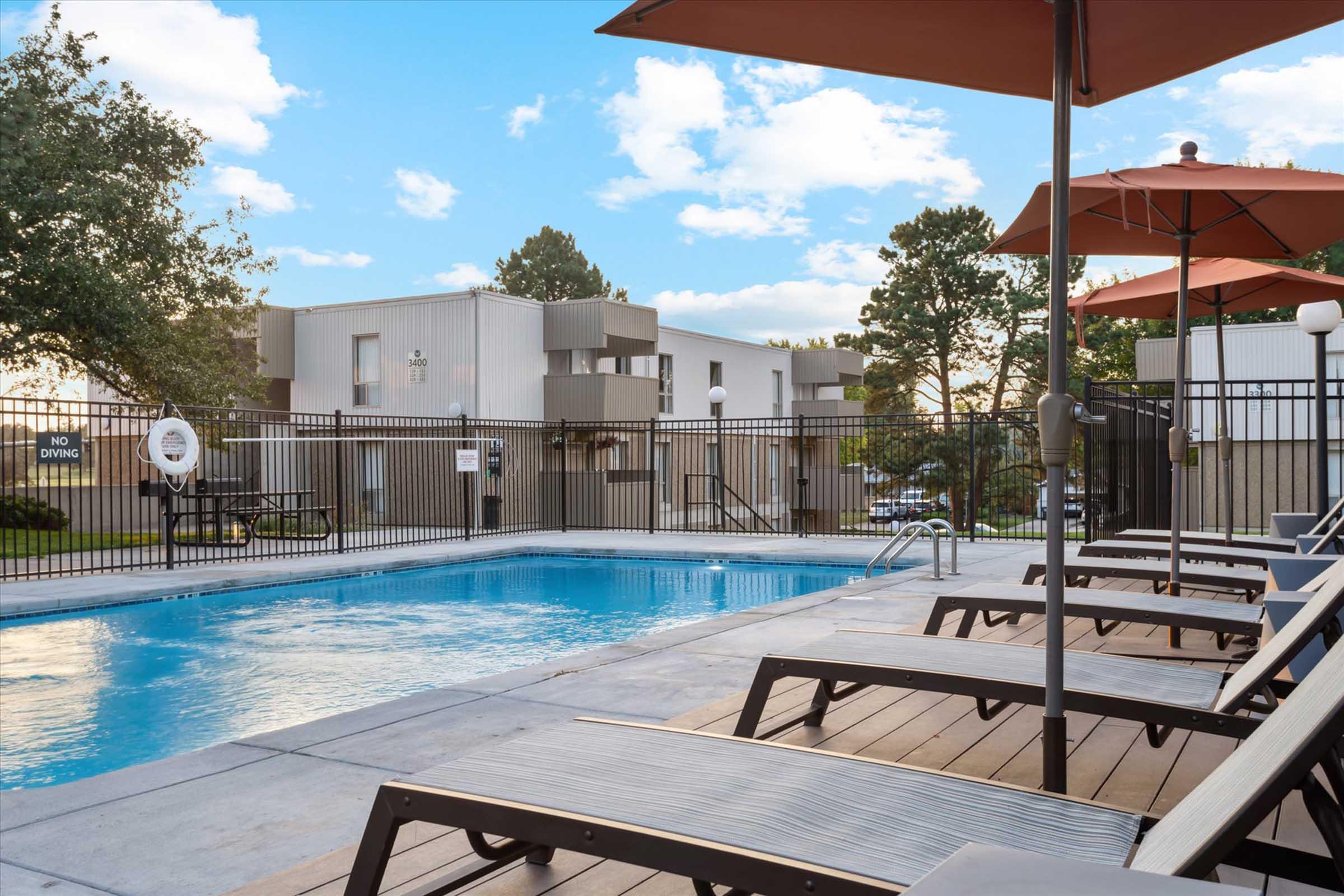 A clear swimming pool surrounded by lounge chairs, with orange umbrellas providing shade. In the background, there are modern apartment buildings set amidst trees and a bright blue sky with scattered clouds. A "No Diving" sign is visible near the pool edge.