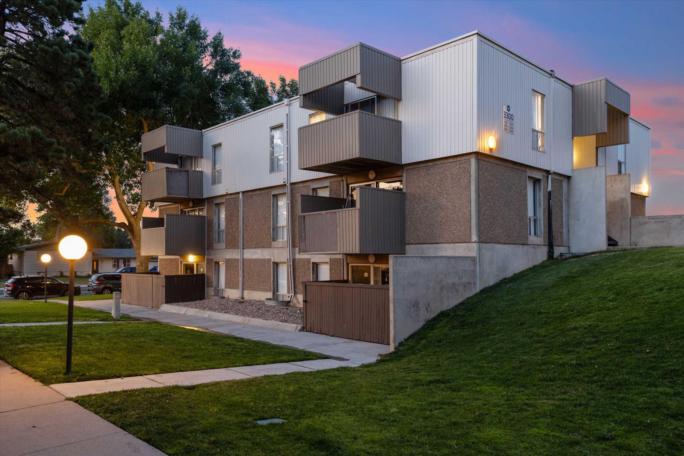 Modern apartment building with multiple balconies and large windows, surrounded by greenery. The exterior features a mix of textured grey walls and sleek metal accents, illuminated by soft outdoor lighting. A manicured lawn in the foreground complements the contemporary architecture against a colorful sunset sky.