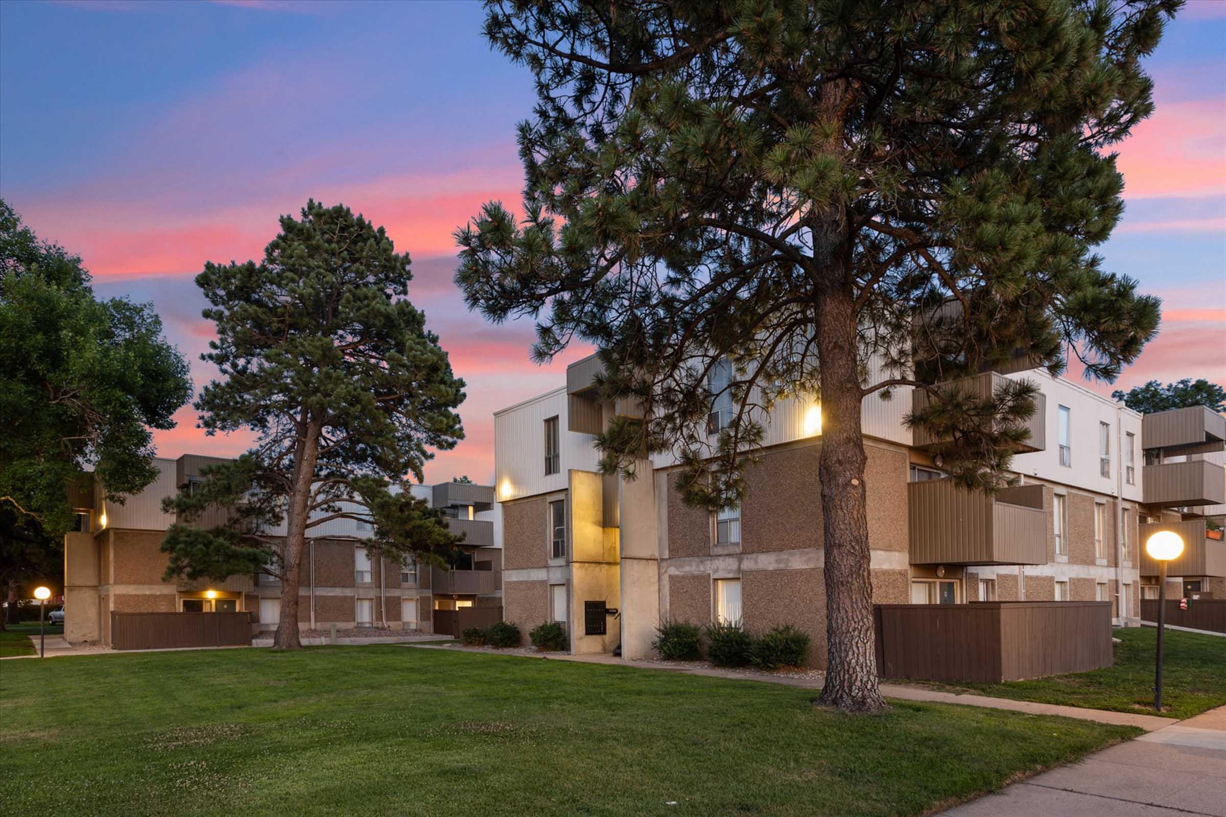 A modern apartment complex with multiple buildings, surrounded by green grass and trees. The scene is illuminated by soft outdoor lighting against a backdrop of a colorful sunset sky, creating a serene atmosphere.