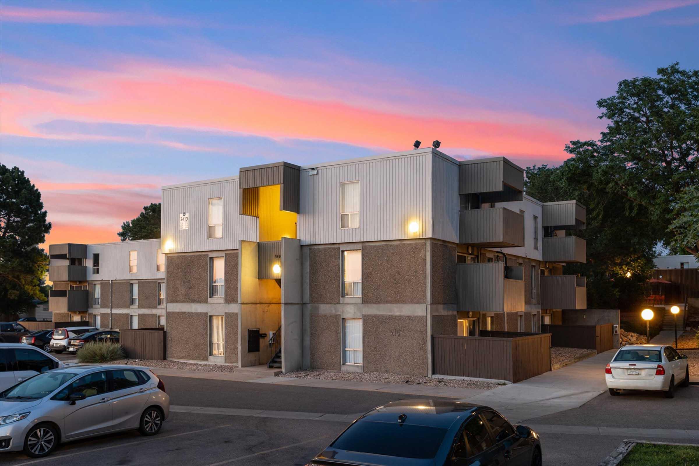 A modern multi-story apartment building with a mix of gray and metallic exterior. The building features balconies and illuminated entryways, set against a colorful sunset sky. Cars are parked in the foreground, and trees can be seen in the surrounding area.