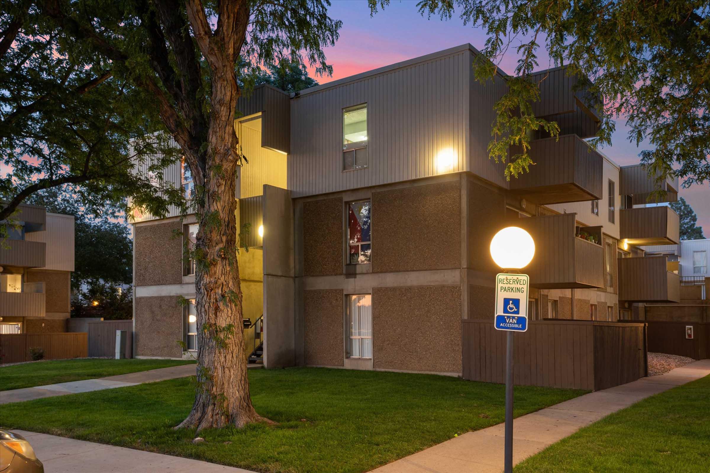 A modern apartment building with a mix of textured gray and metal exterior, surrounded by lush green grass and trees. The structure features large windows and balcony spaces. A lighted sign indicates accessible parking nearby. The scene is set during twilight, with a soft pink sky in the background.