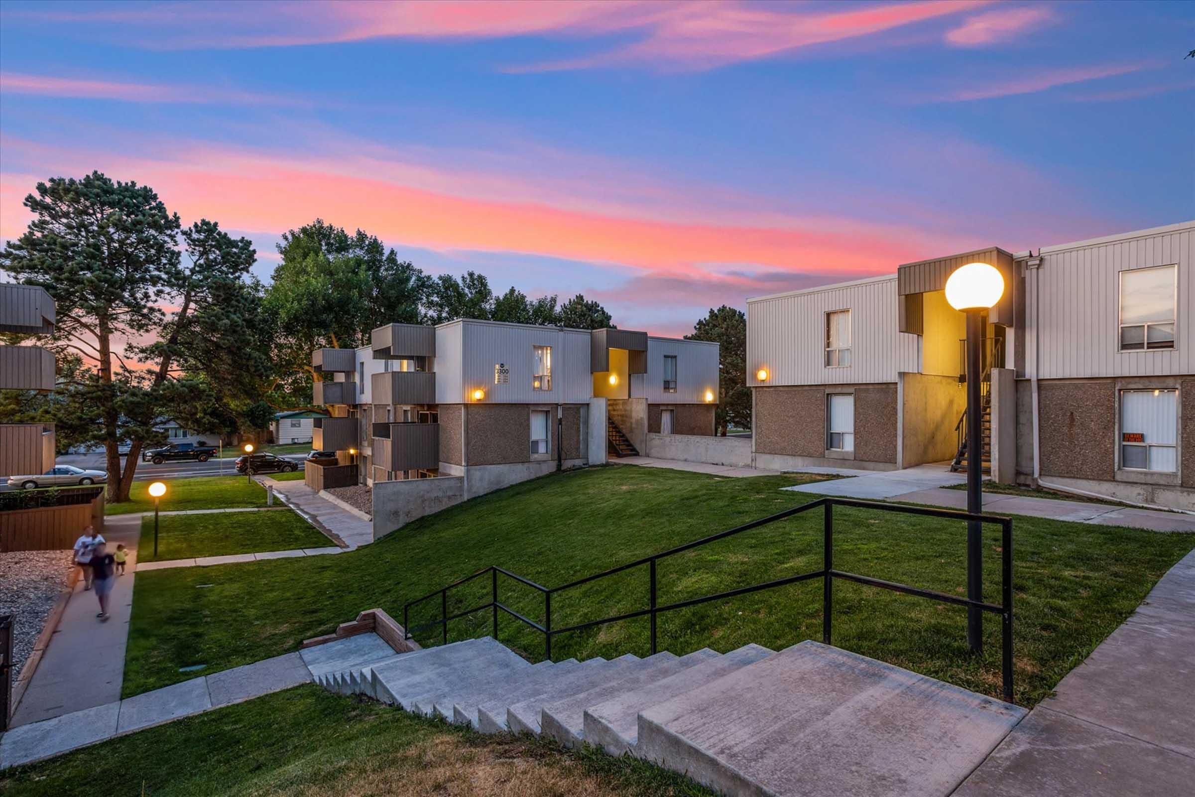 A landscaped apartment complex during sunset, featuring beige and gray buildings with illuminated windows. A grassy area is in the foreground, with steps leading up to the buildings and a walking path. The sky displays vibrant hues of pink and purple, creating a serene atmosphere. Trees are present in the background.