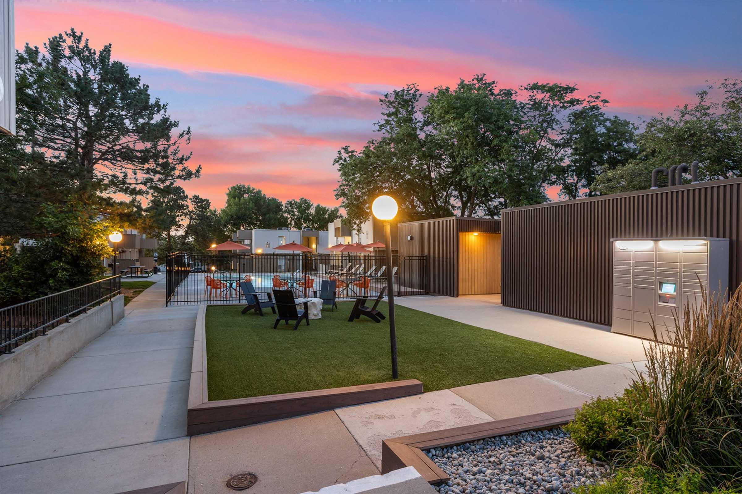 A tranquil outdoor space during sunset, featuring a grassy area with lounge chairs and a table, surrounded by trees. Soft lighting from a lamp and a modern mailbox structure complete the scene, creating a serene atmosphere perfect for relaxation.